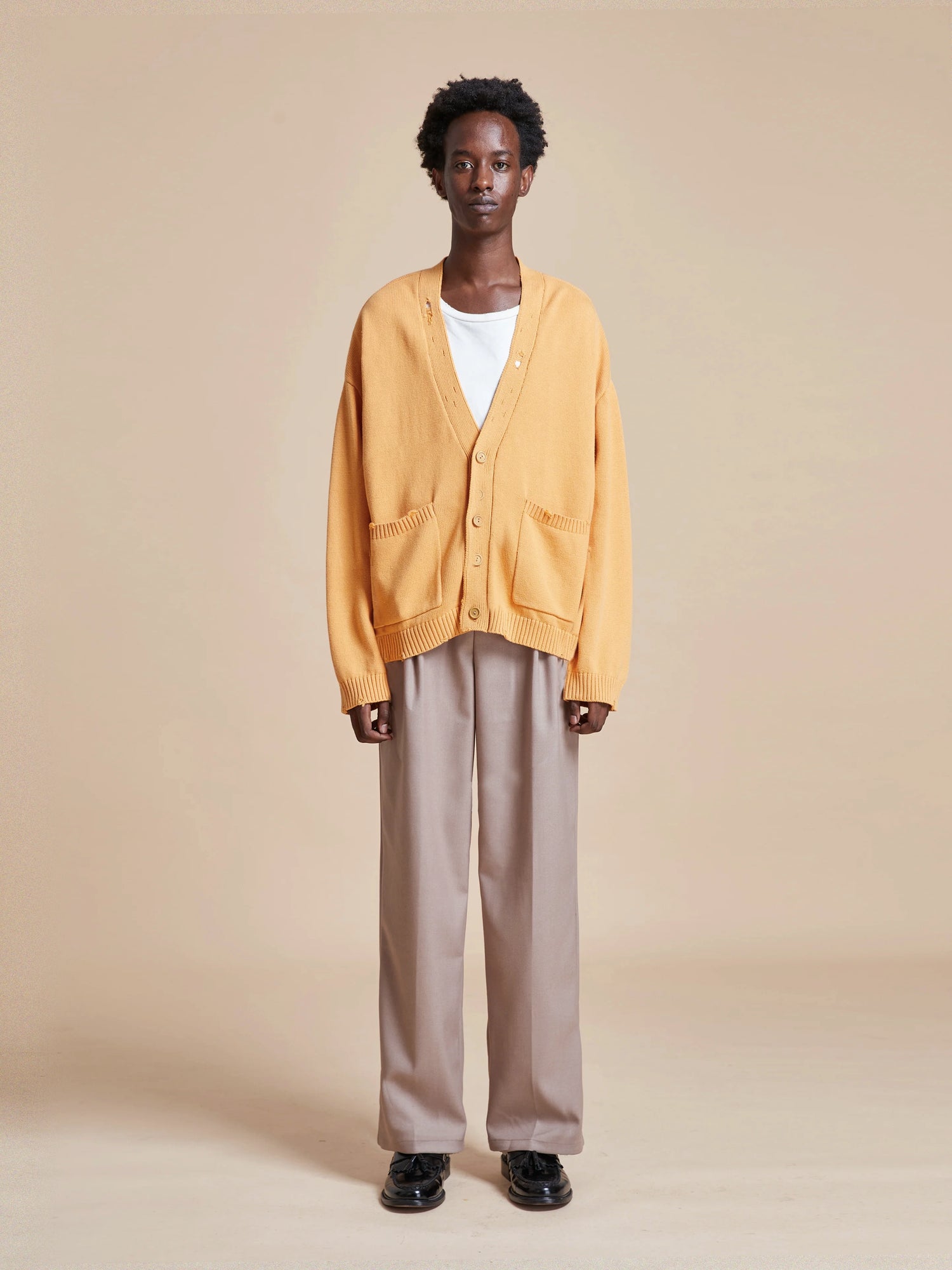A man wearing a Found Cadmium Distressed Cardigan sweater and tan trousers.