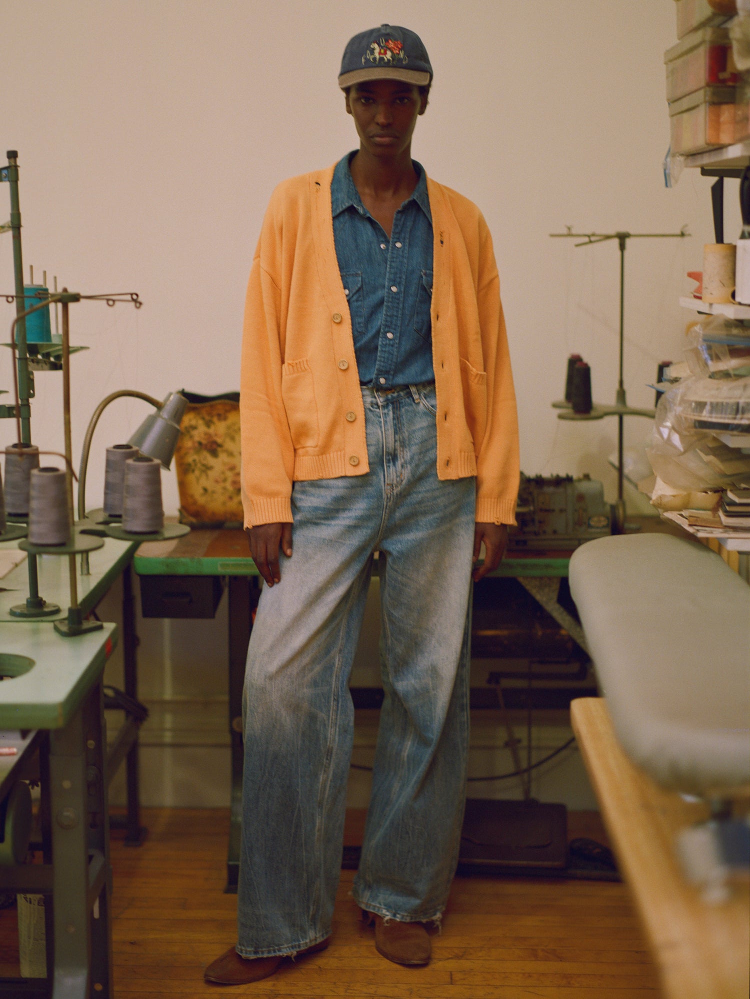 A man in jeans and a hat, wearing a Cadmium Distressed Cardigan sweater by Found, standing in a workshop.