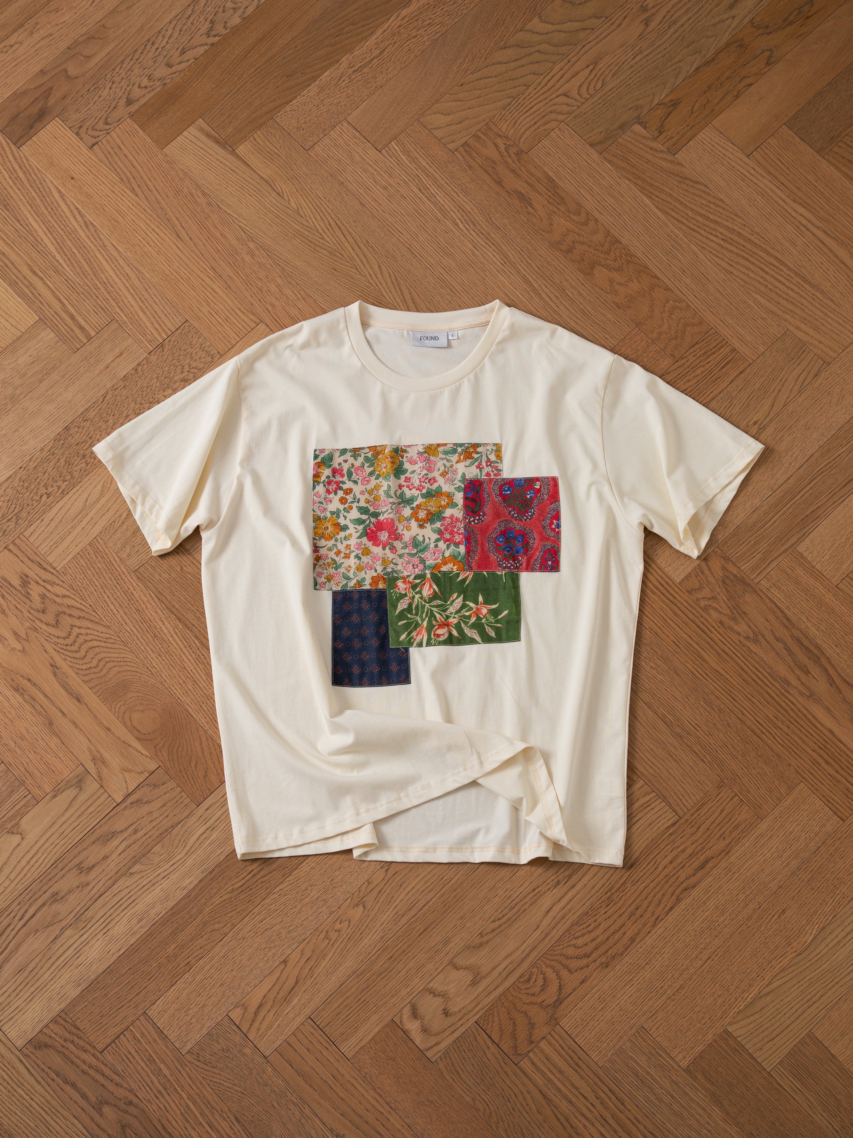 A beige Patchwork Tee by FOUND, featuring four colorful patchwork squares with various floral patterns, laid flat on a wooden herringbone floor.