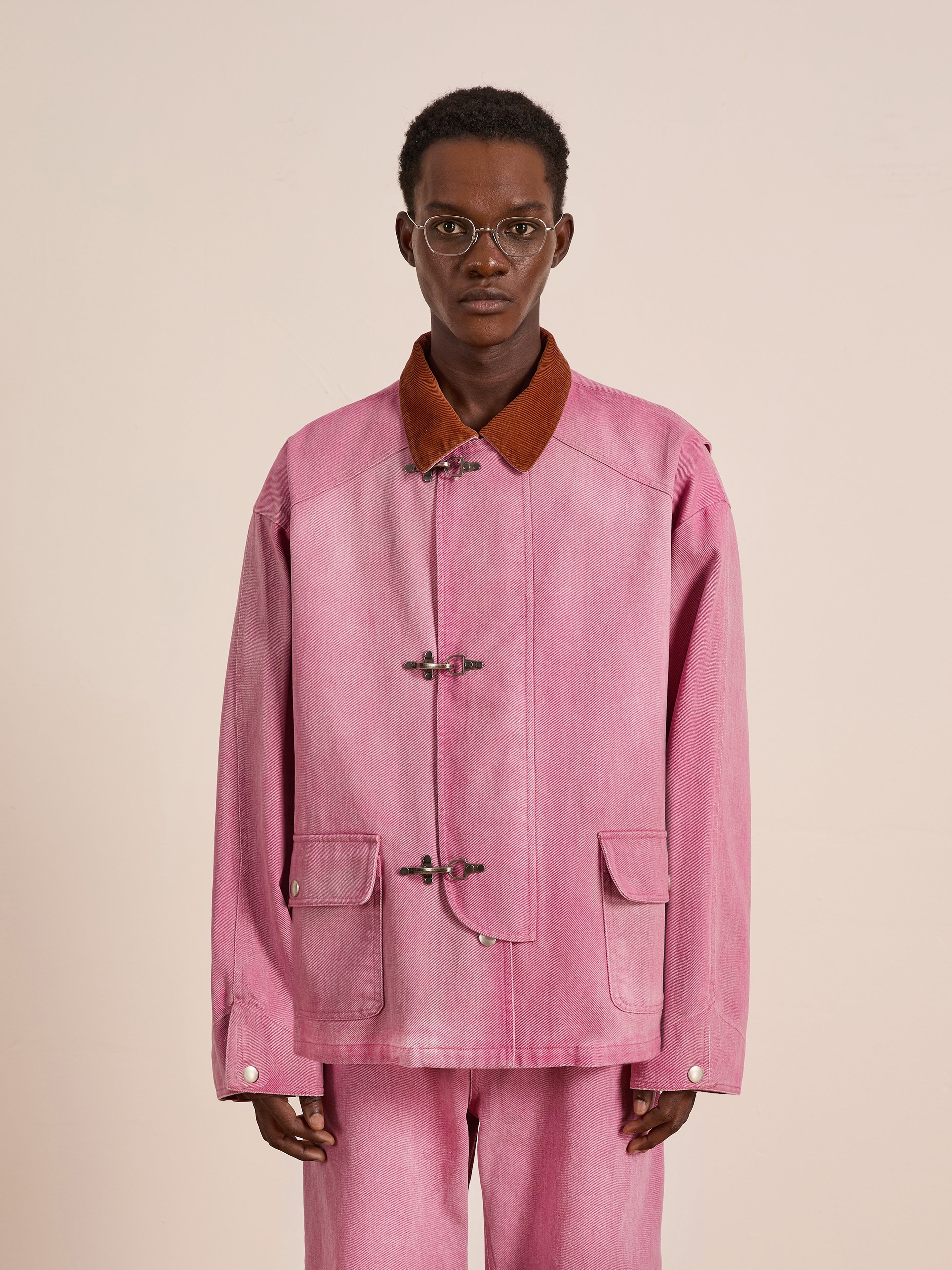 A person wearing glasses models the Found Washed Fireman Clasp Workshop Jacket in a loose pink fit with matching pants, standing against a plain light background.