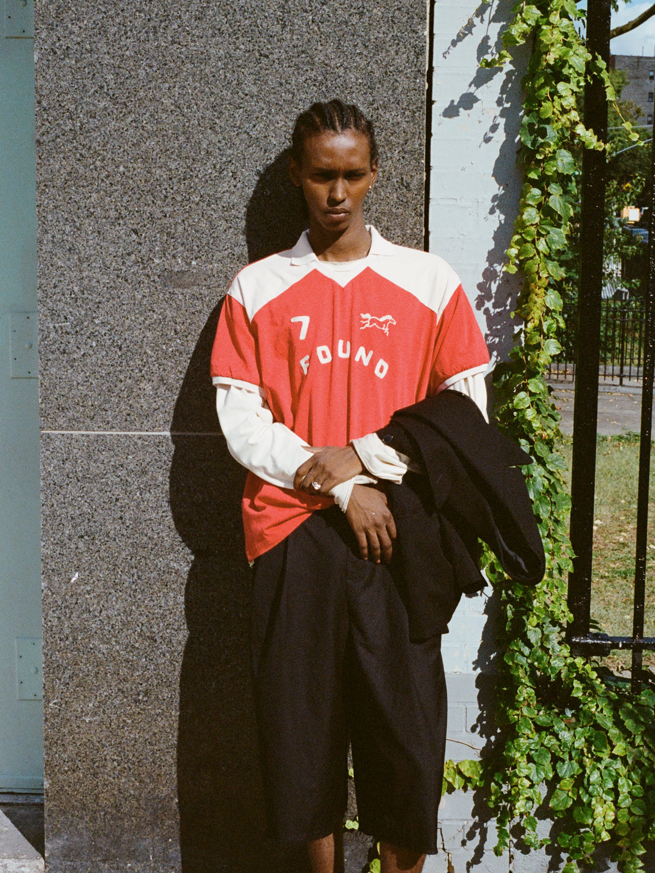 A person with braided hair stands against a wall wearing a Goji Red Jersey by FOUND, styled like vintage athletic uniforms with the number 7 and "FOUND" on it. They hold a black jacket as green vines frame the right side of the scene.