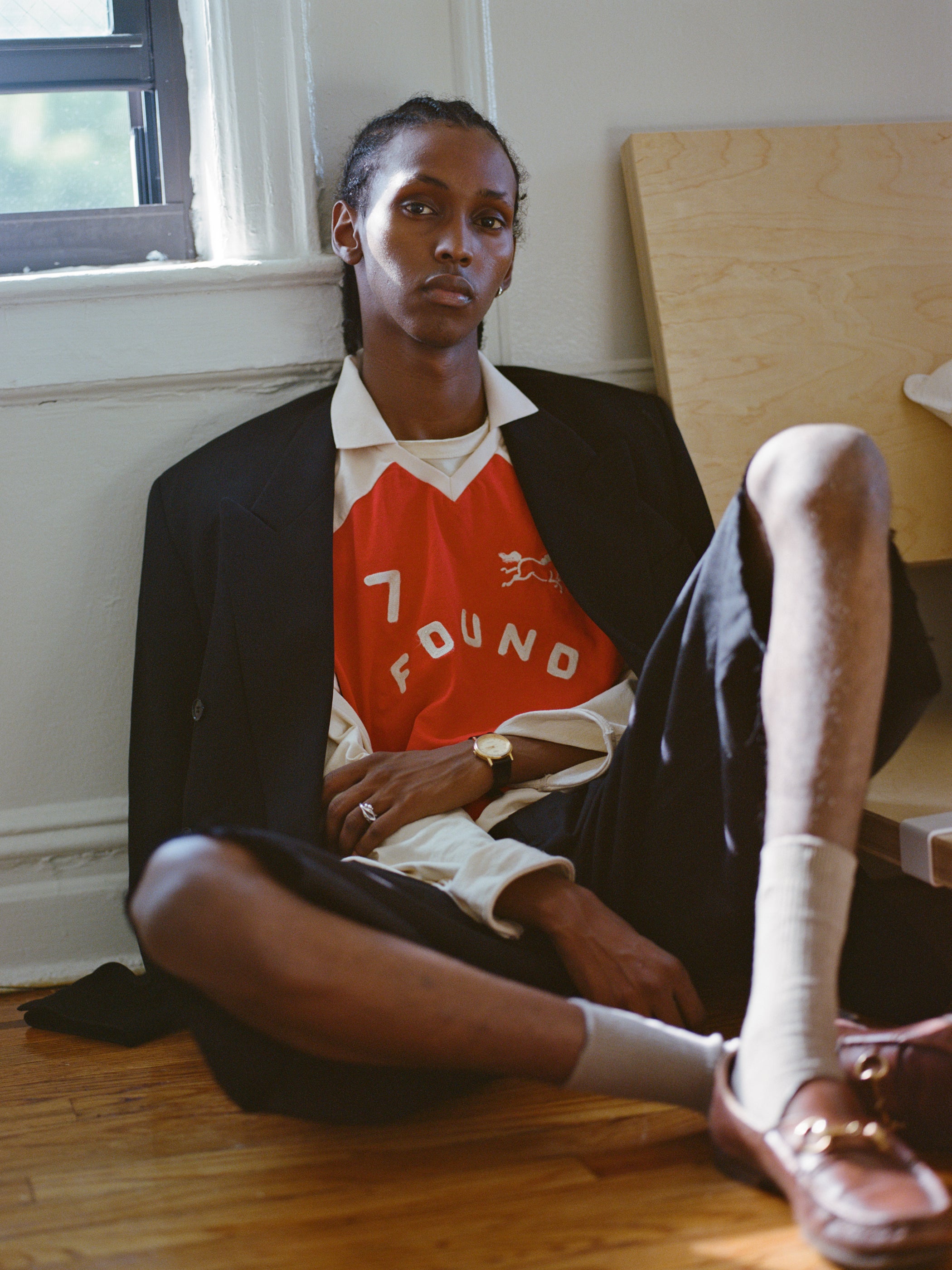 Sitting on a wooden floor near a window, the person wears FOUND's Goji Red Jersey, paired with a black jacket, shorts, and brown shoes while leaning against a wall.