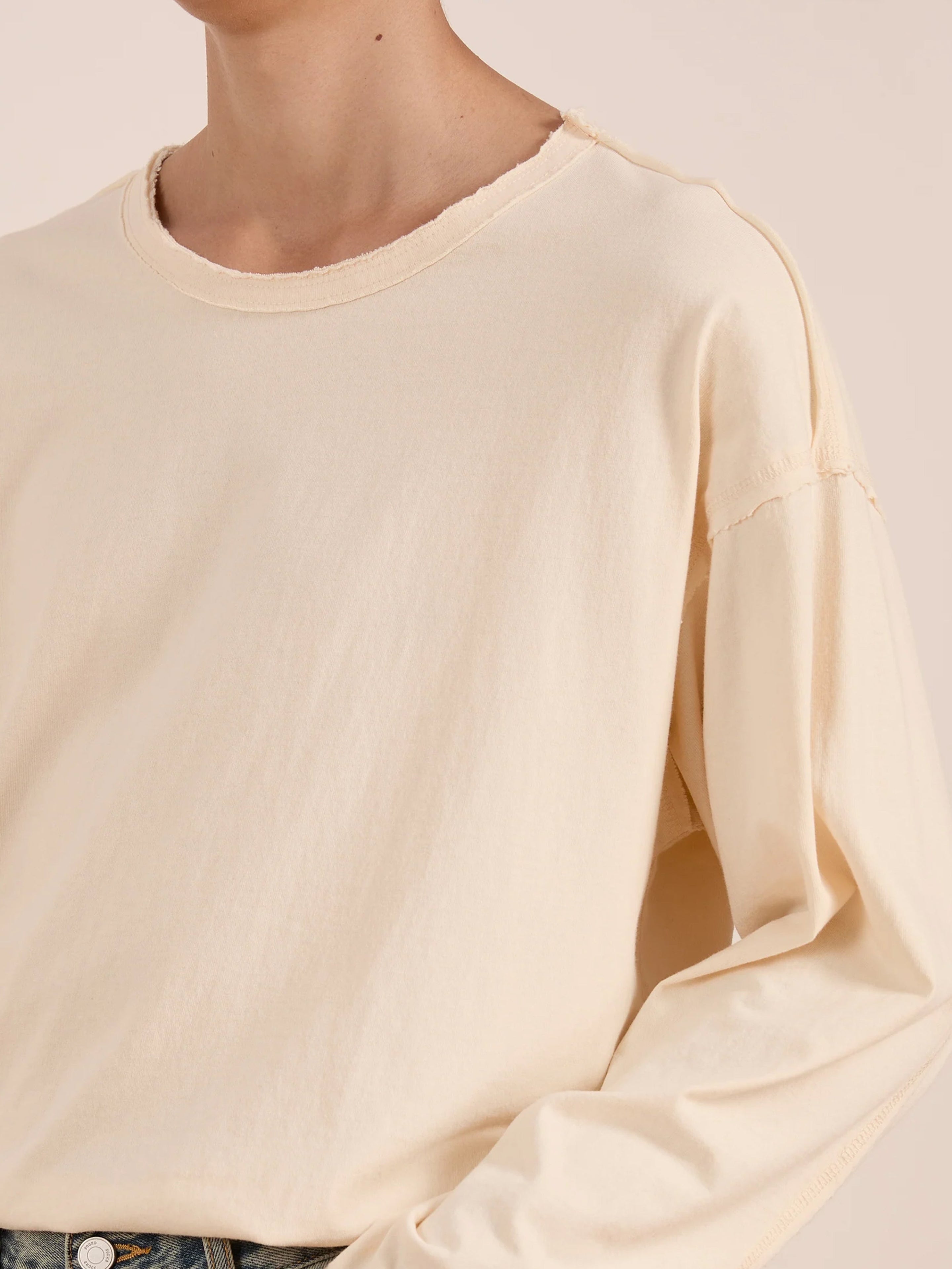 A person wearing the FOUND Reversed LS Tee, a unisex off-white long sleeve top featuring exposed seams and a distressed neckline, shown from shoulders to waist.