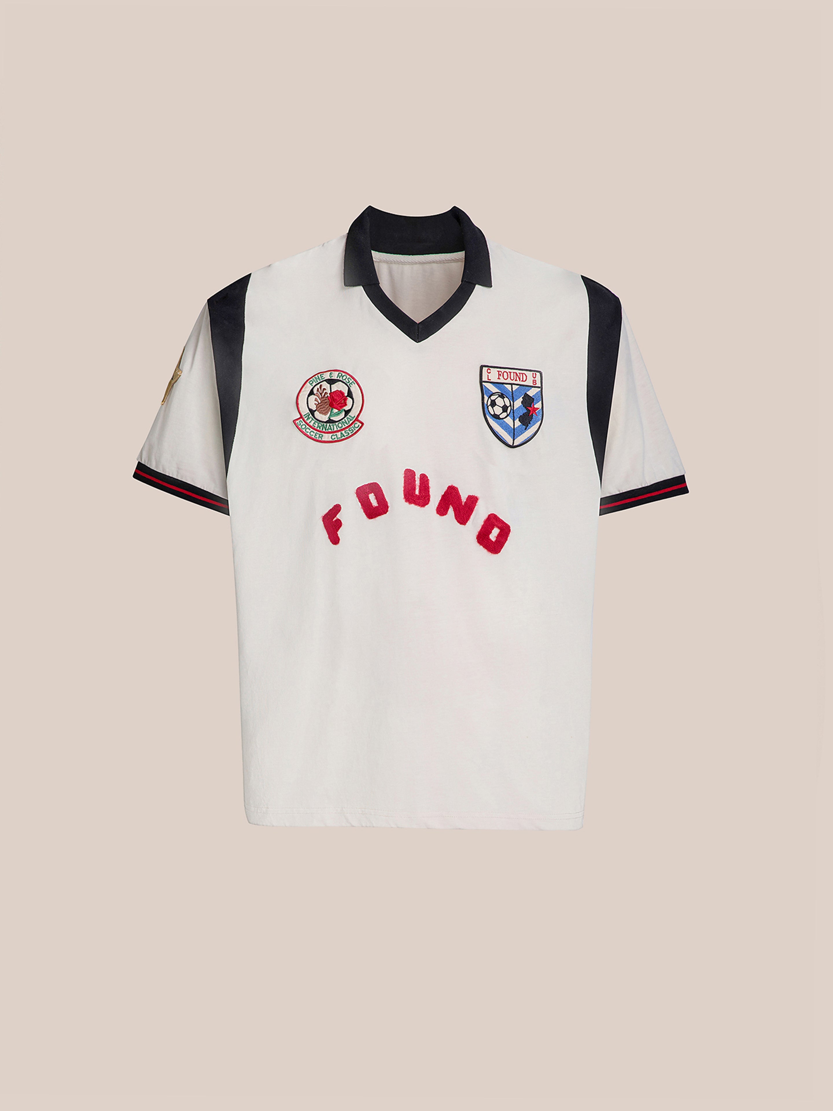 The Union Soccer Jersey by FOUND is a white jersey with a black collar and sleeves, featuring embroidered logos and "FOUND" in red across the front, offering a contemporary sportswear piece with vintage football vibes for those who love classic appeal.