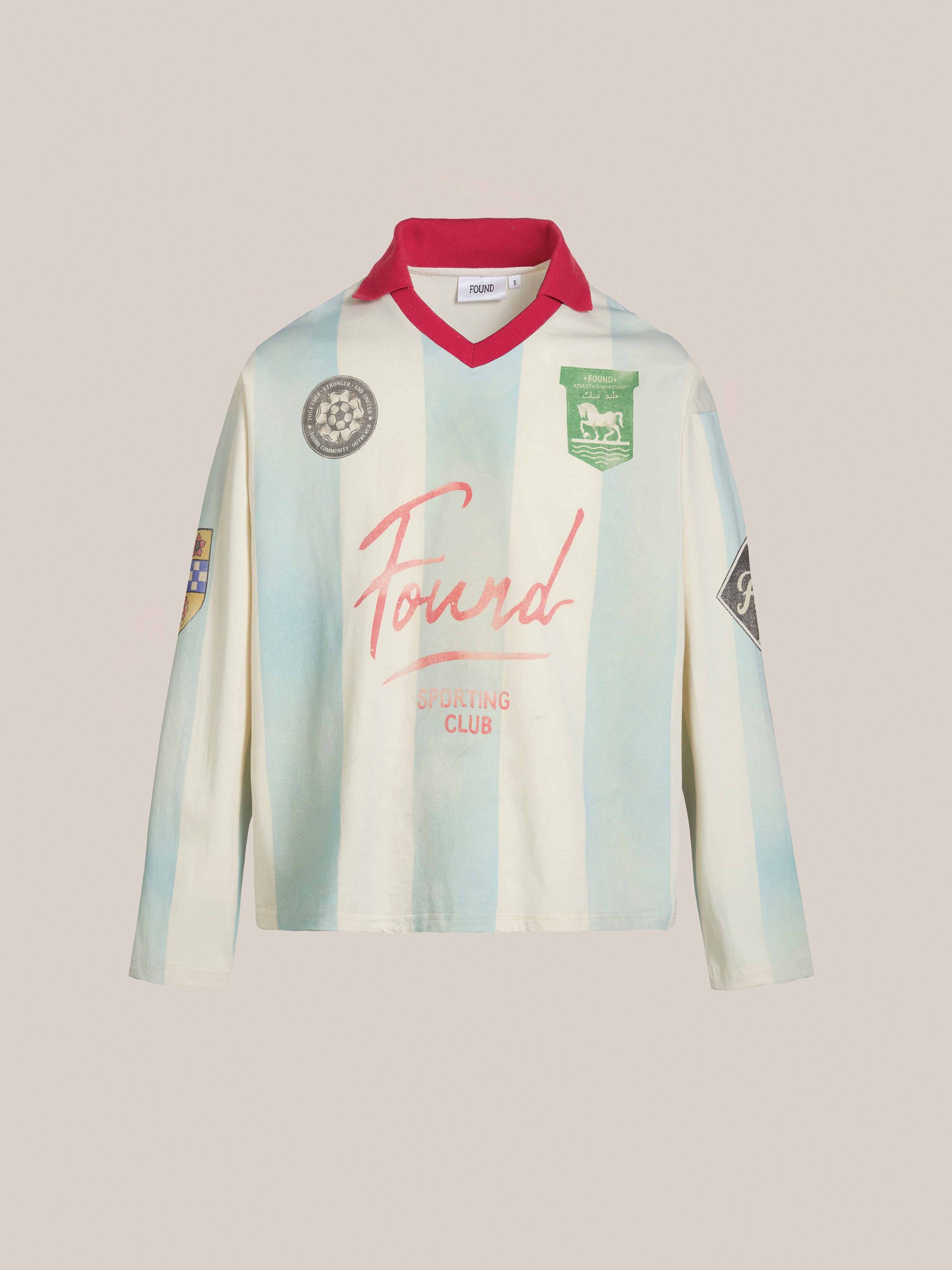 The Stripe Sport LS Jersey by FOUND features light blue and white vertical stripes, a red collar, and various patches/logos. This heritage-inspired sportswear piece offers a unisex relaxed fit that merges past aesthetics with modern style.