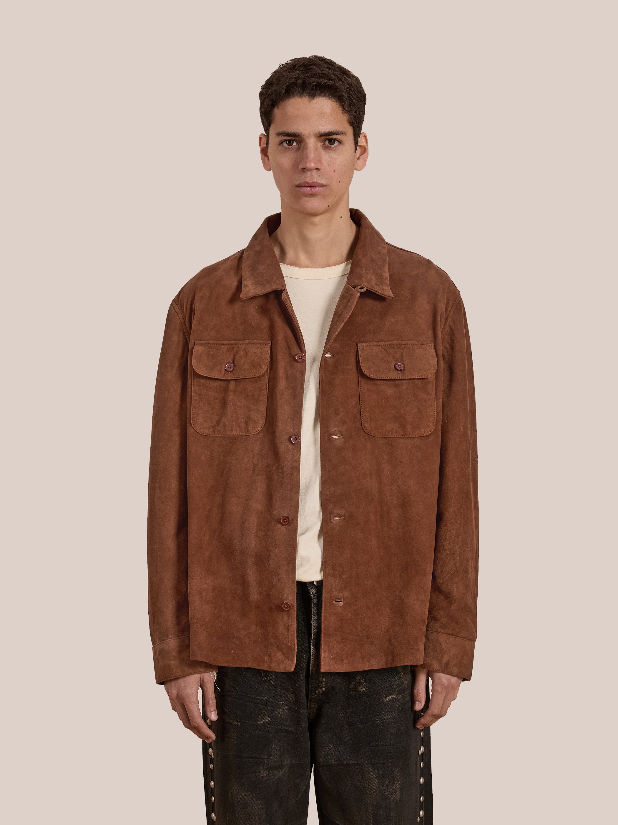 A person faces forward, wearing the FOUND Suede Leather Camp Shirt—a unisex relaxed fit goat suede shirt with two chest pockets—layered over a beige shirt and paired with dark pants.