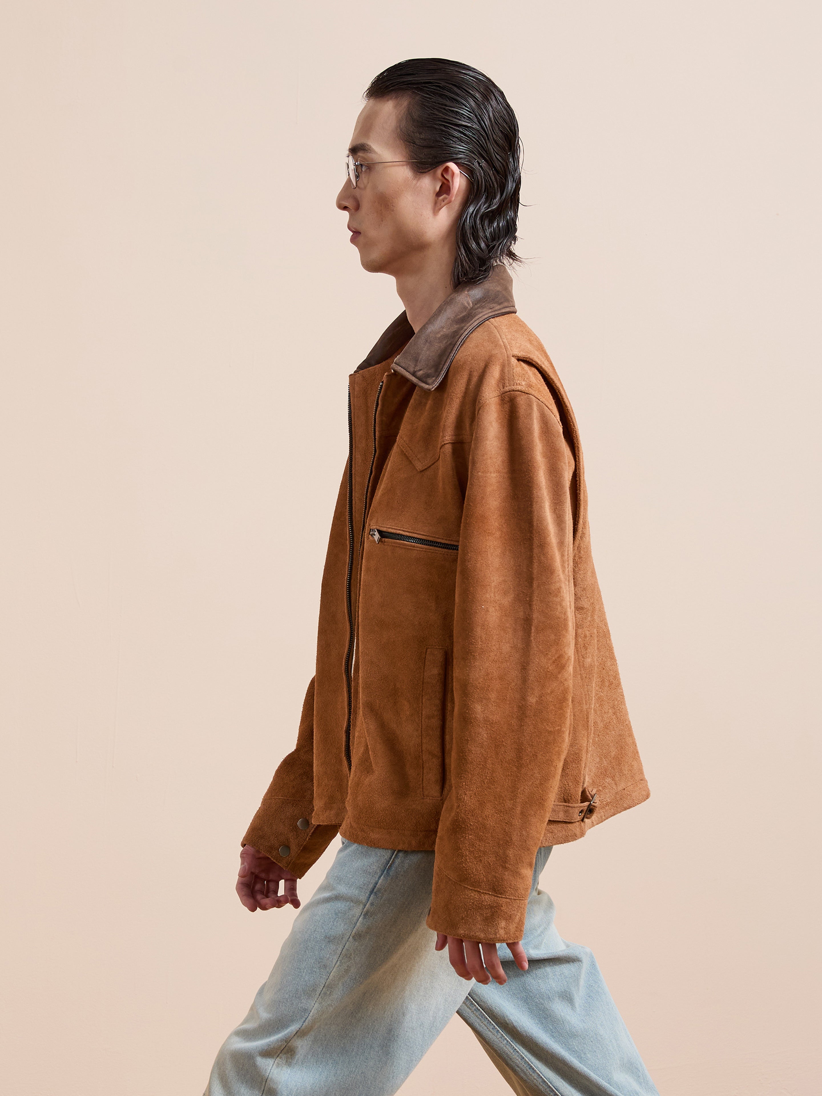 A person with slicked-back hair and glasses wears the FOUND Suede Western Barn Jacket and light blue jeans while walking against a plain beige background.