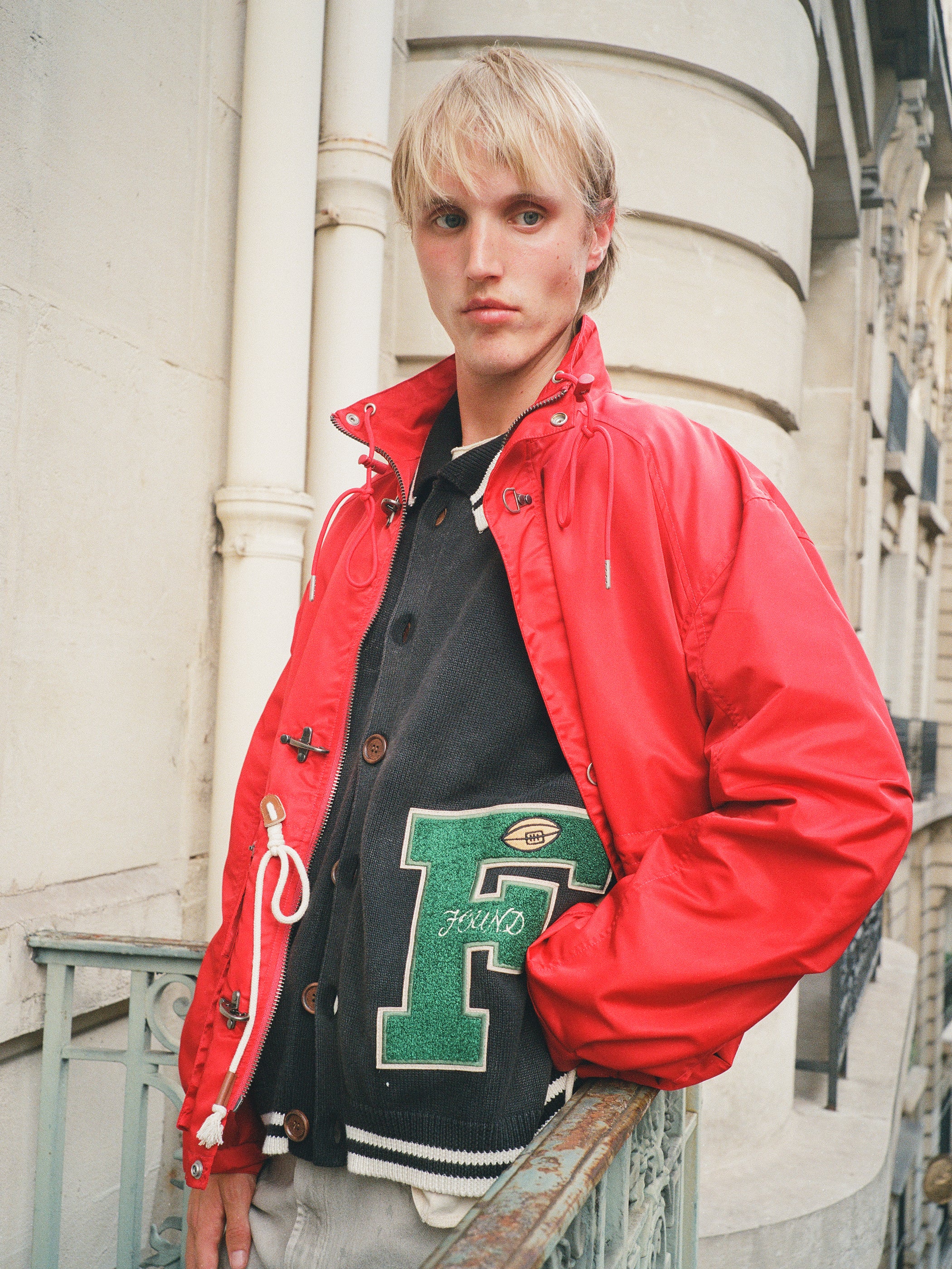 A person with blond hair stands on a balcony, wearing a red jacket over the FOUND Varsity Patch Collared Cardigan featuring a bold chenille F patch.