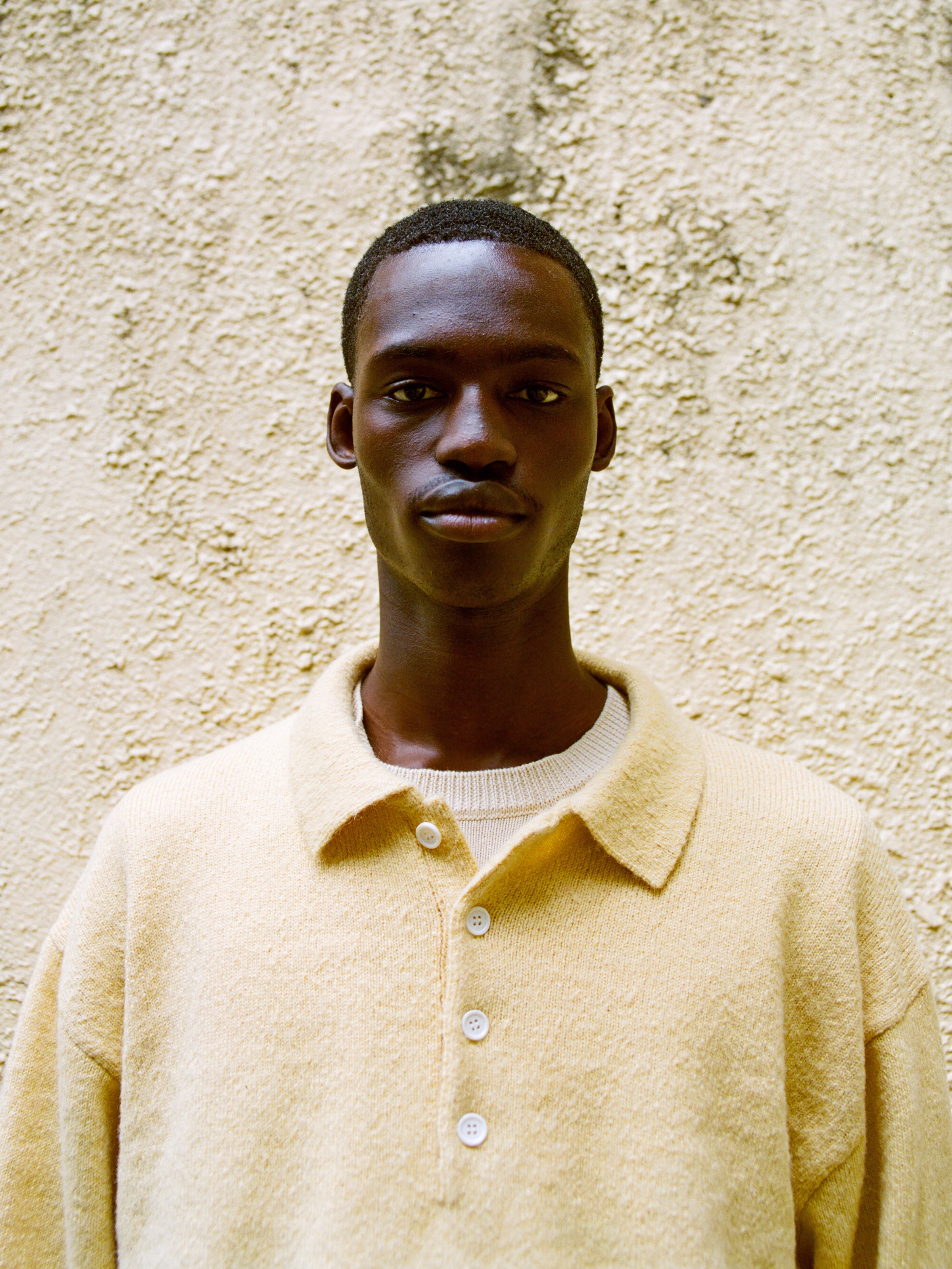 A man in a beige FOUND Knit LS Polo Sweater, made from brushed knit fabric, stands before a textured yellow wall and looks directly at the camera.