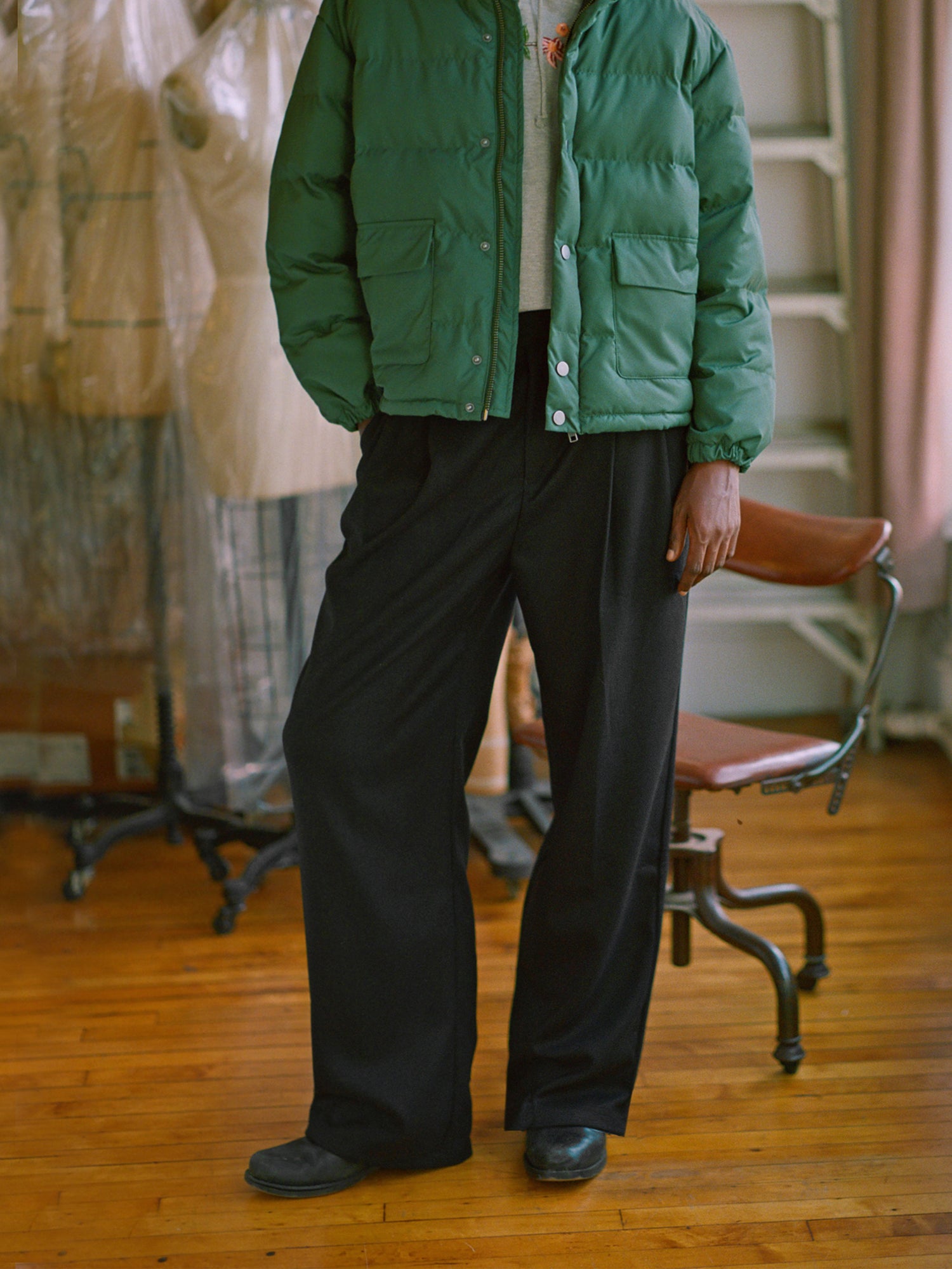 A woman in a green puffer jacket standing in a room, showcasing the pleated trousers from Found, featuring the classic silhouette.