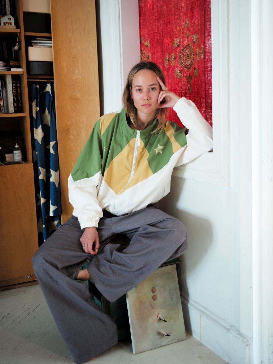 A person in a FOUND Legacy Cricket Track Jacket sits on a small stool beside a painting, with a red and gold patterned curtain in the background. The vintage athletic gear complements their gray pants, adding an intriguing contrast to the artistic setting.