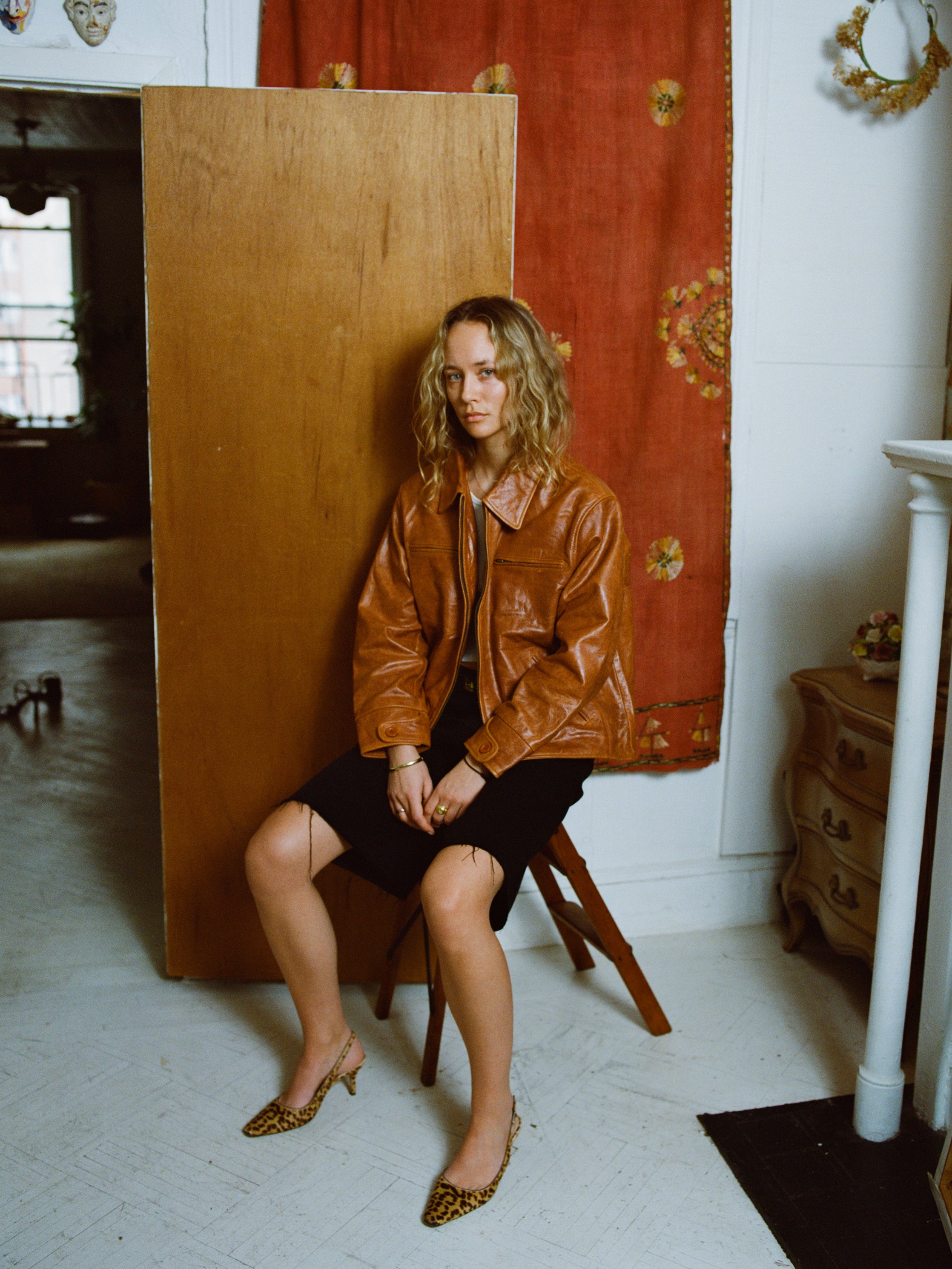 Seated elegantly on a wooden chair, a woman with curly hair showcases the FOUND Marlboro Sun Faded Leather Racer Jacket, paired stylishly with a black skirt and leopard print shoes, reflecting her dedication to sustainable fashion through ethically sourced materials.