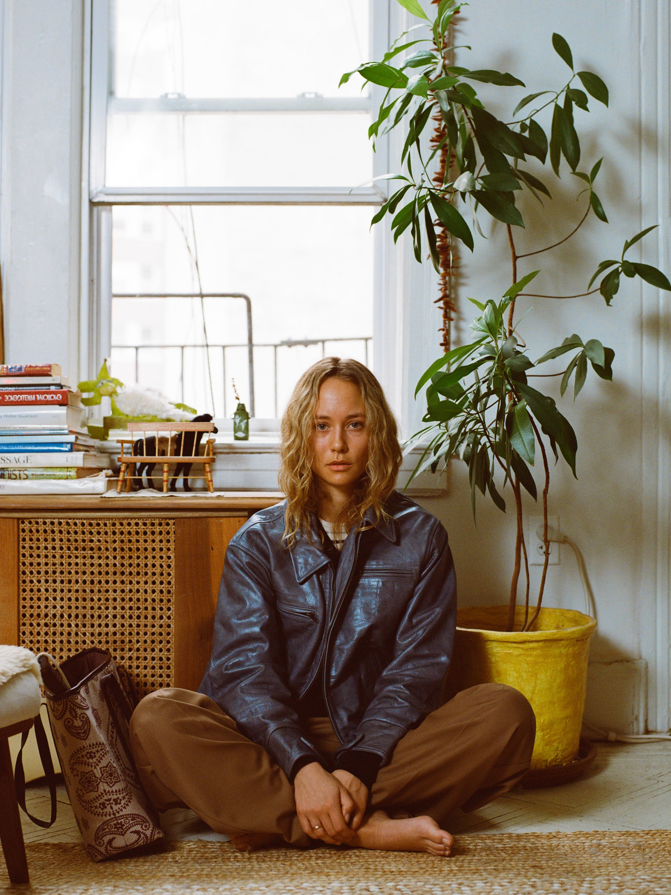 A person sits cross-legged on a woven rug by a window, wearing a FOUND Marlboro Sun Faded Leather Racer Jacket and brown pants. Beside them is a tall potted plant, with a stack of books on the left.