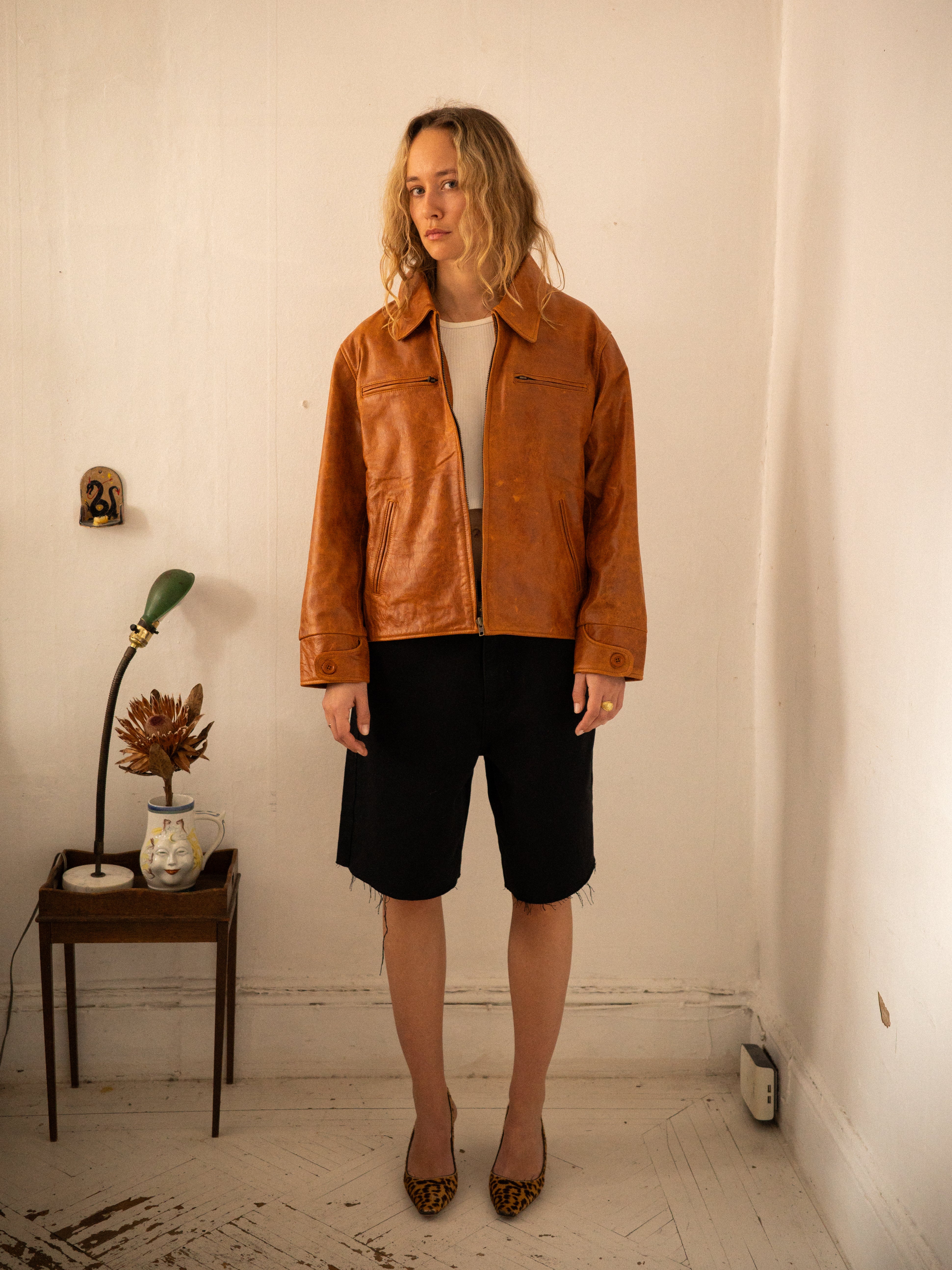 A person with long hair wears a FOUND Marlboro Sun Faded Leather Racer Jacket, black shorts, and patterned flats while standing indoors near a small table with a lamp and vase.