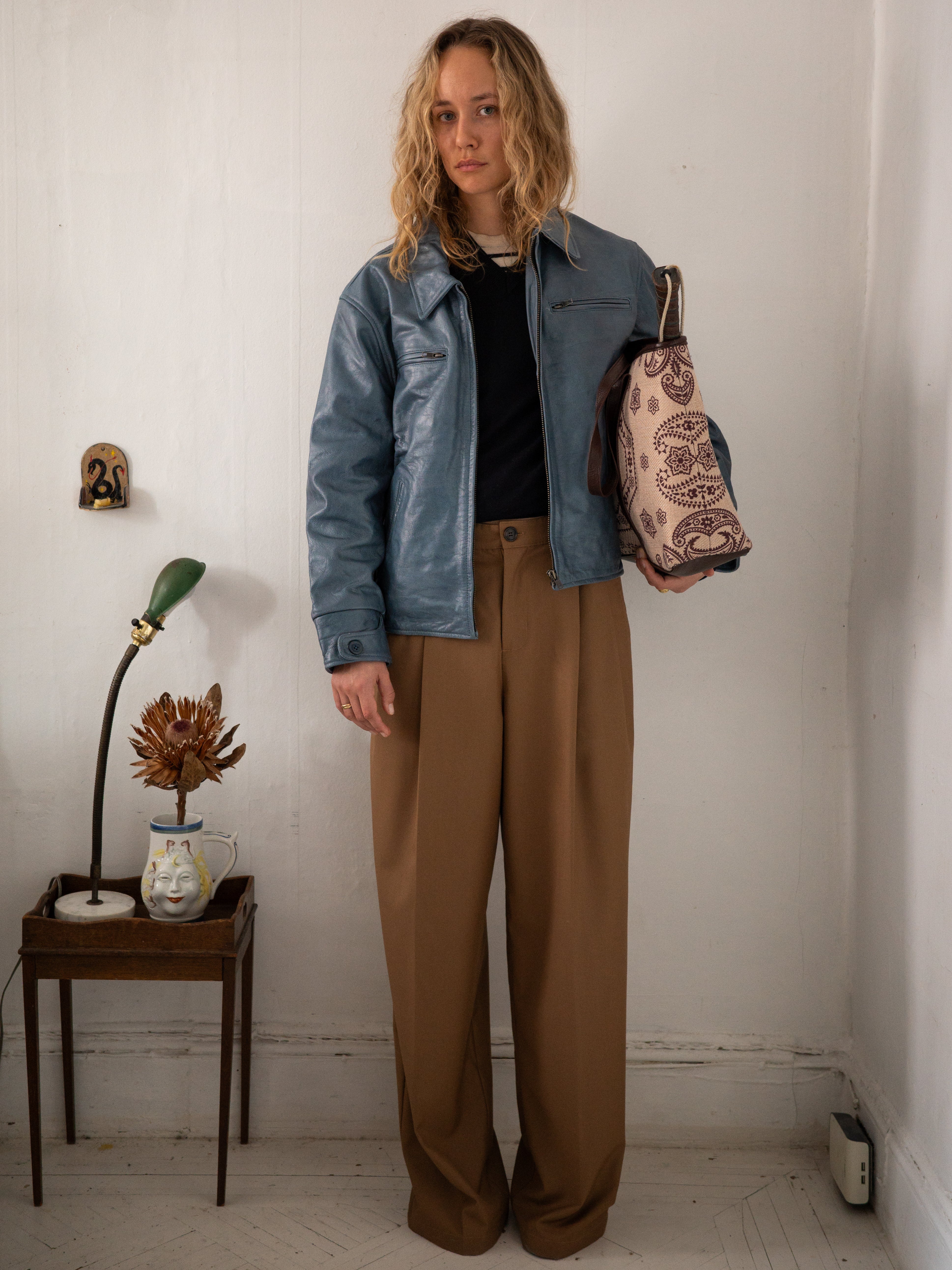 A person with long blonde hair stands in a room wearing a FOUND Marlboro Sun Faded Leather Racer Jacket, a black top, and brown trousers, holding a light pink bag. A small table with a flower and lamp is beside them.