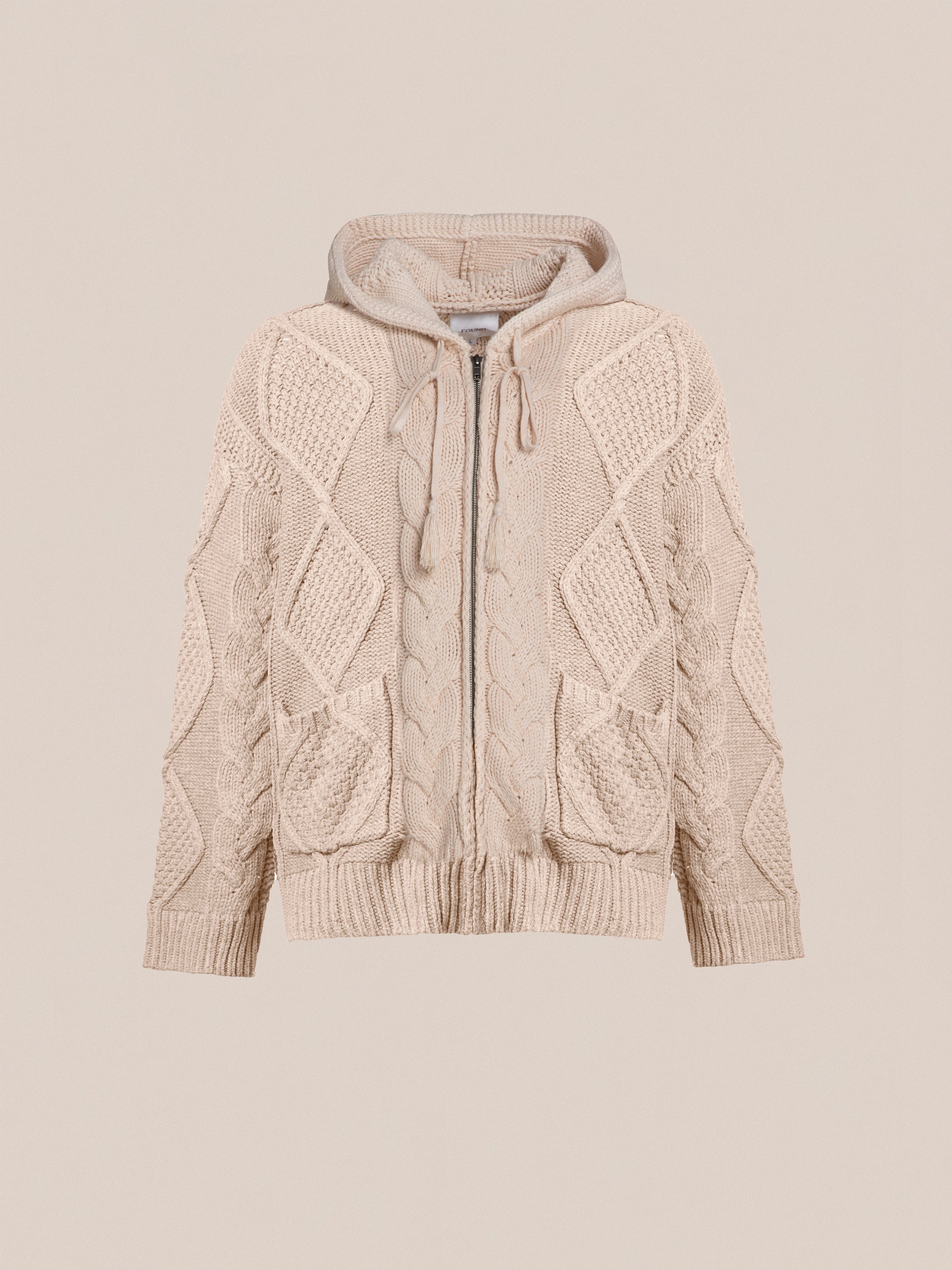 A beige, chunky knit, zip-up hoodie with a hood, featuring intricate cable knit patterns and two front pockets for comfort and warmth is the Zip Up Cable Knit Hoodie by Found.