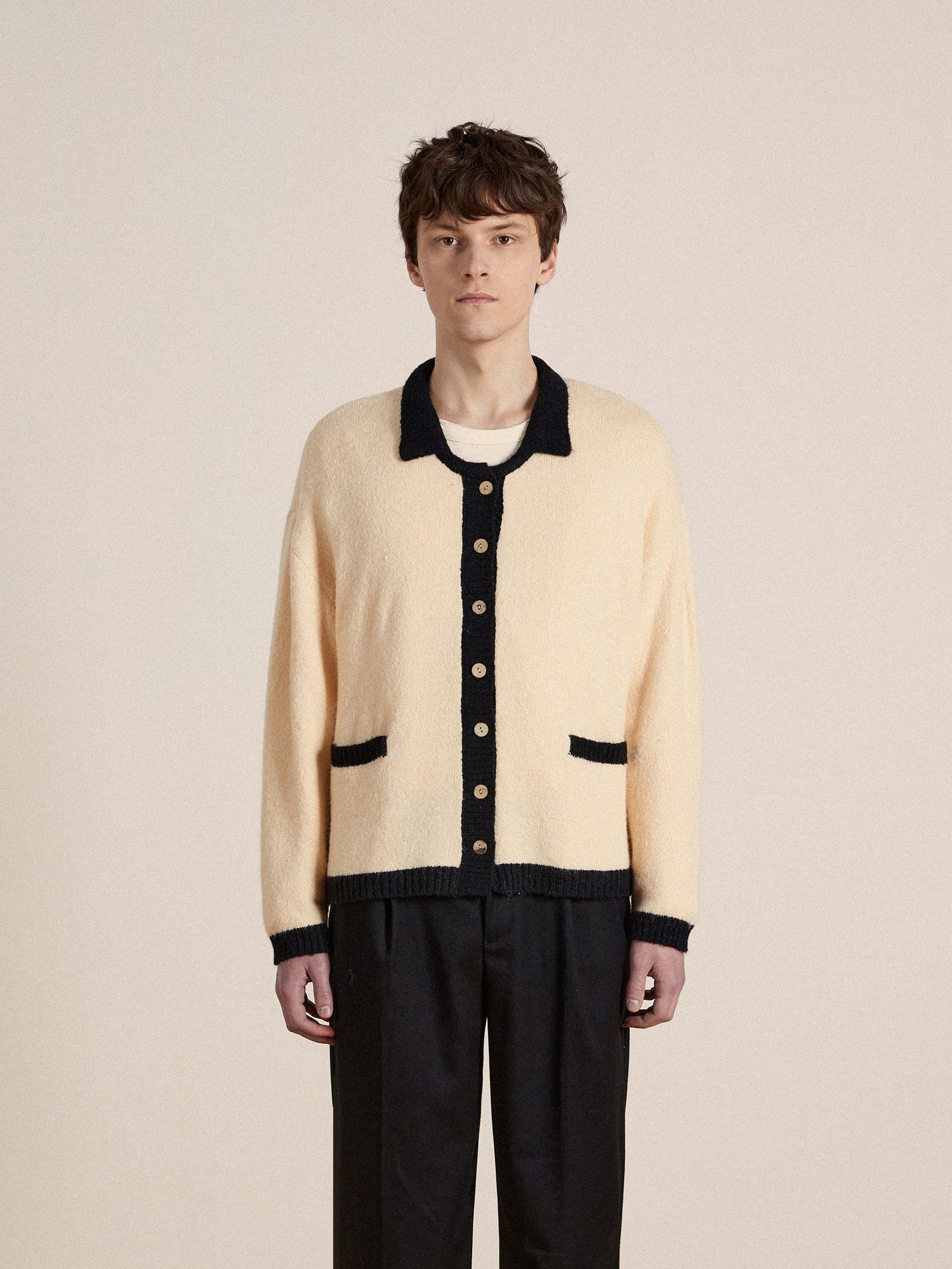 A man wearing a cream Found Sima Contrast Collar Knitted Cardigan and black trousers.