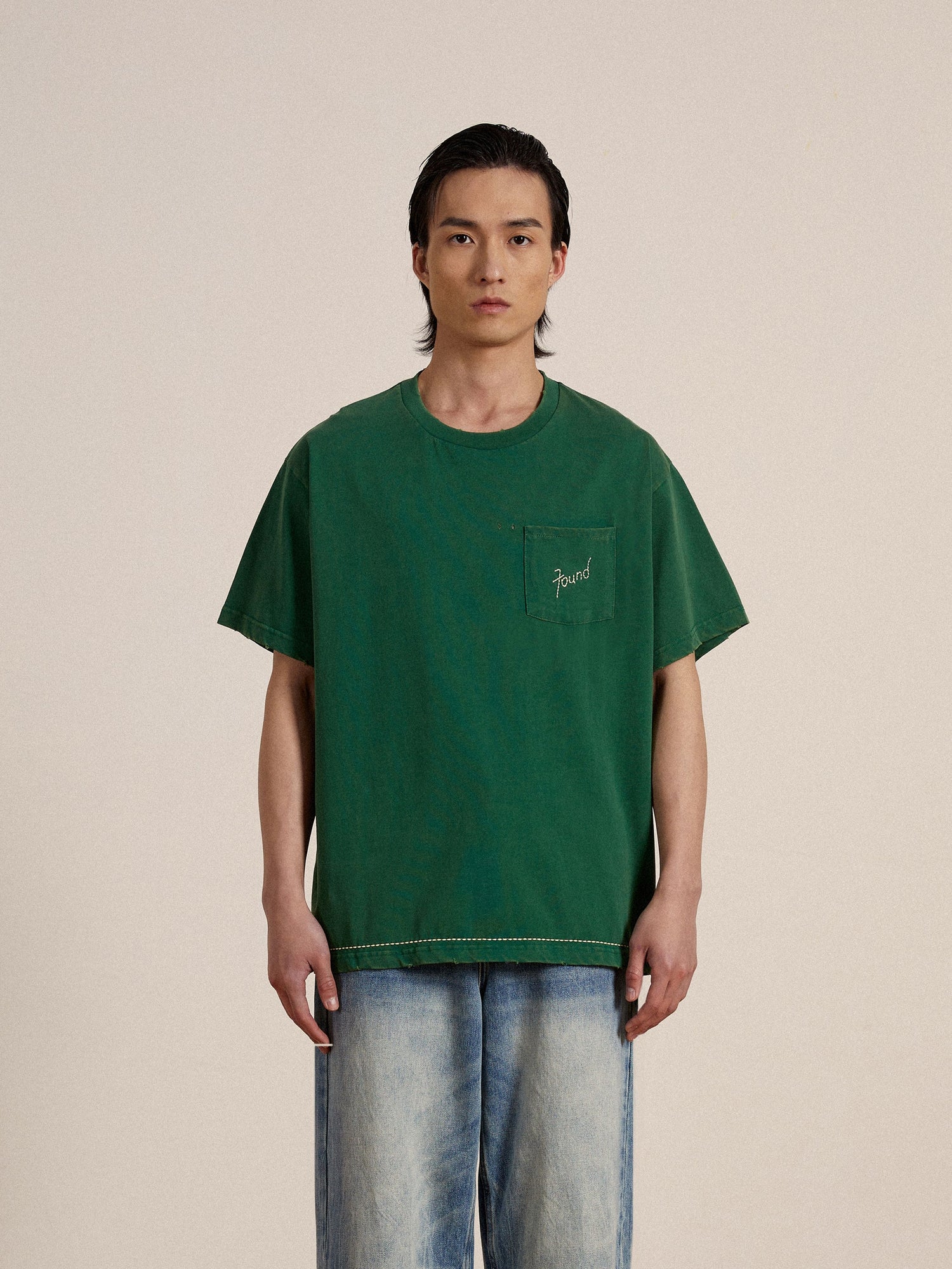 A man wearing a green Found embroidered logo tee and jeans with distressing.