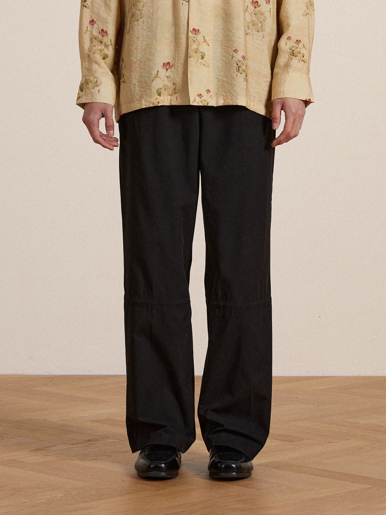 A person wearing a floral-patterned yellow shirt and black Found Tencel pleated pants, standing with hands slightly behind them, focusing on the lower half of the body.