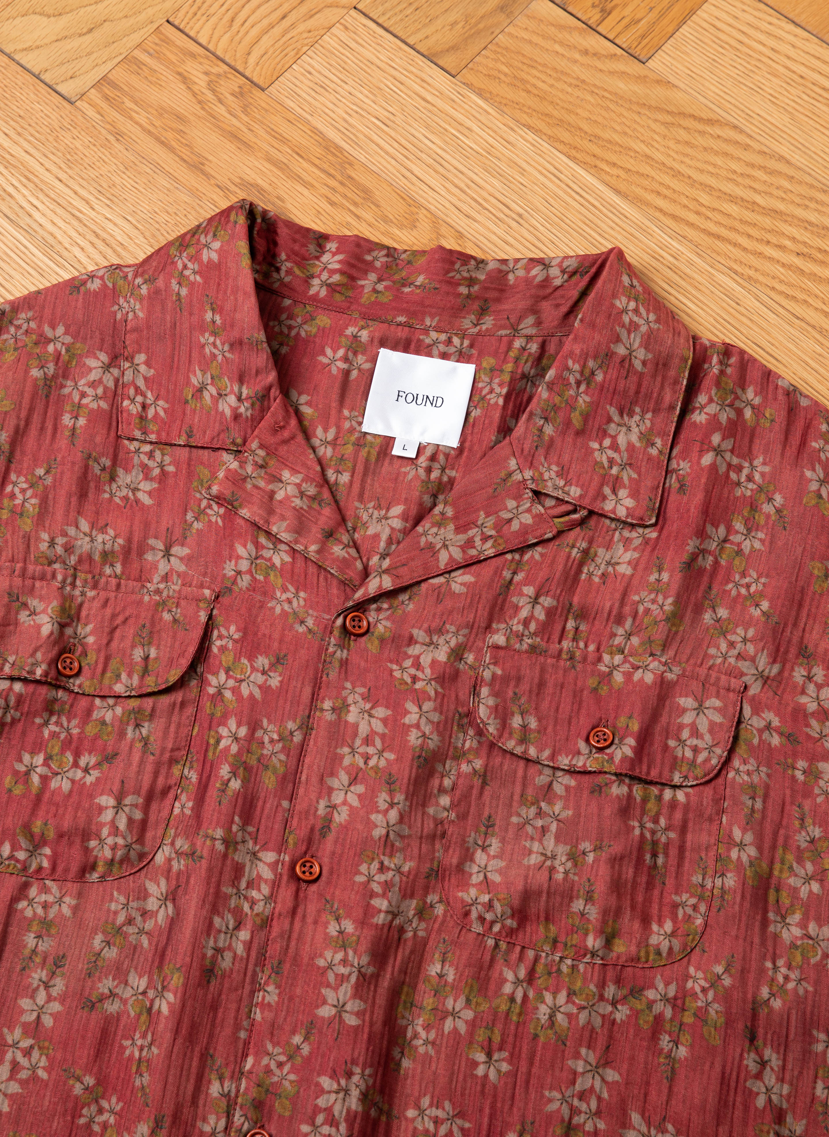 A red floral-patterned short-sleeve shirt with a "FOUND" label lies flat on a wooden floor. The shirt, reminiscent of the Charlot LS Camp Shirt, features dual chest pockets with buttons.