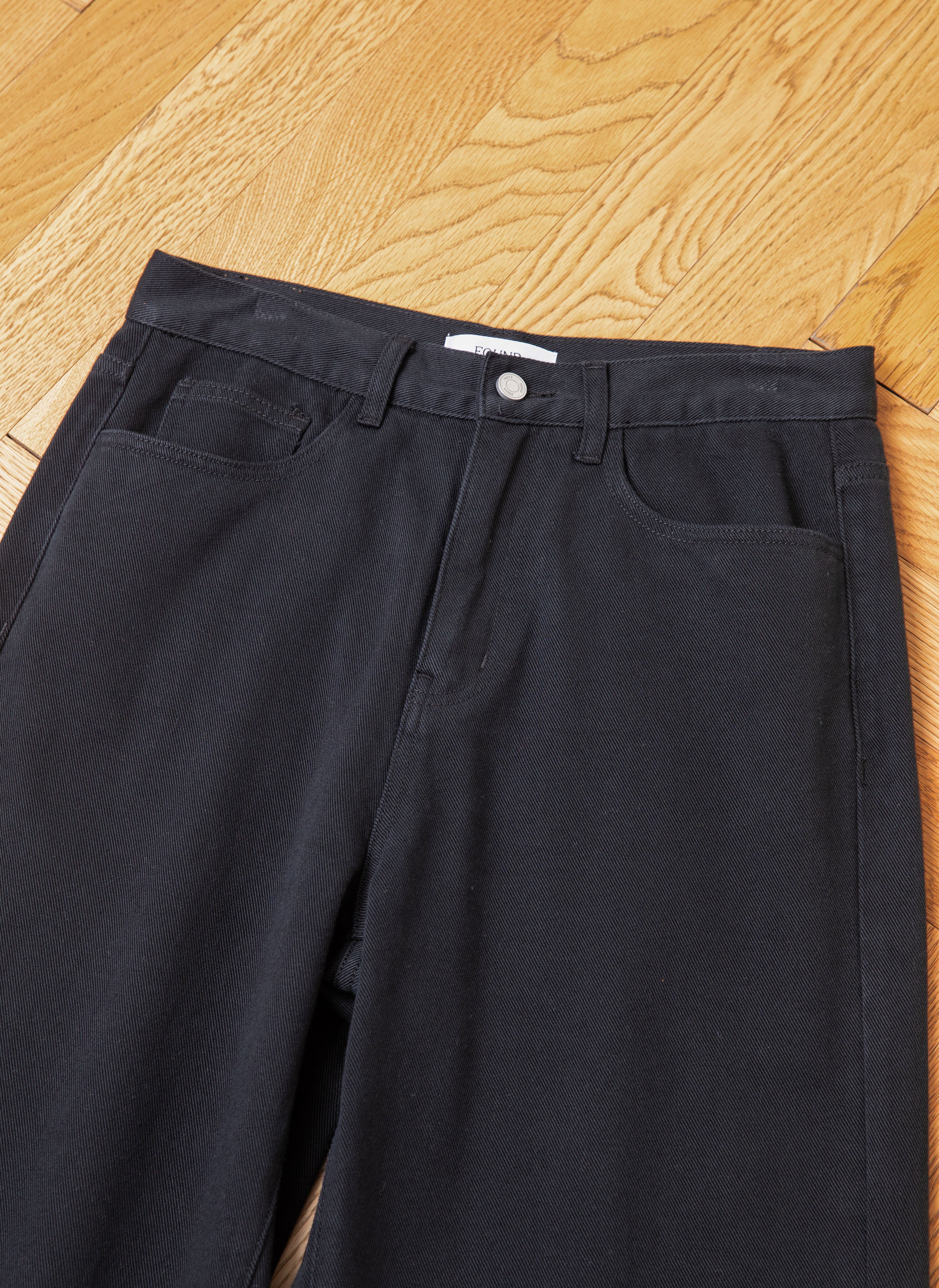 A pair of black wash, mid rise Lacy Baggy Jeans by FOUND with a metal button and zip fly, laid flat on a wooden floor.