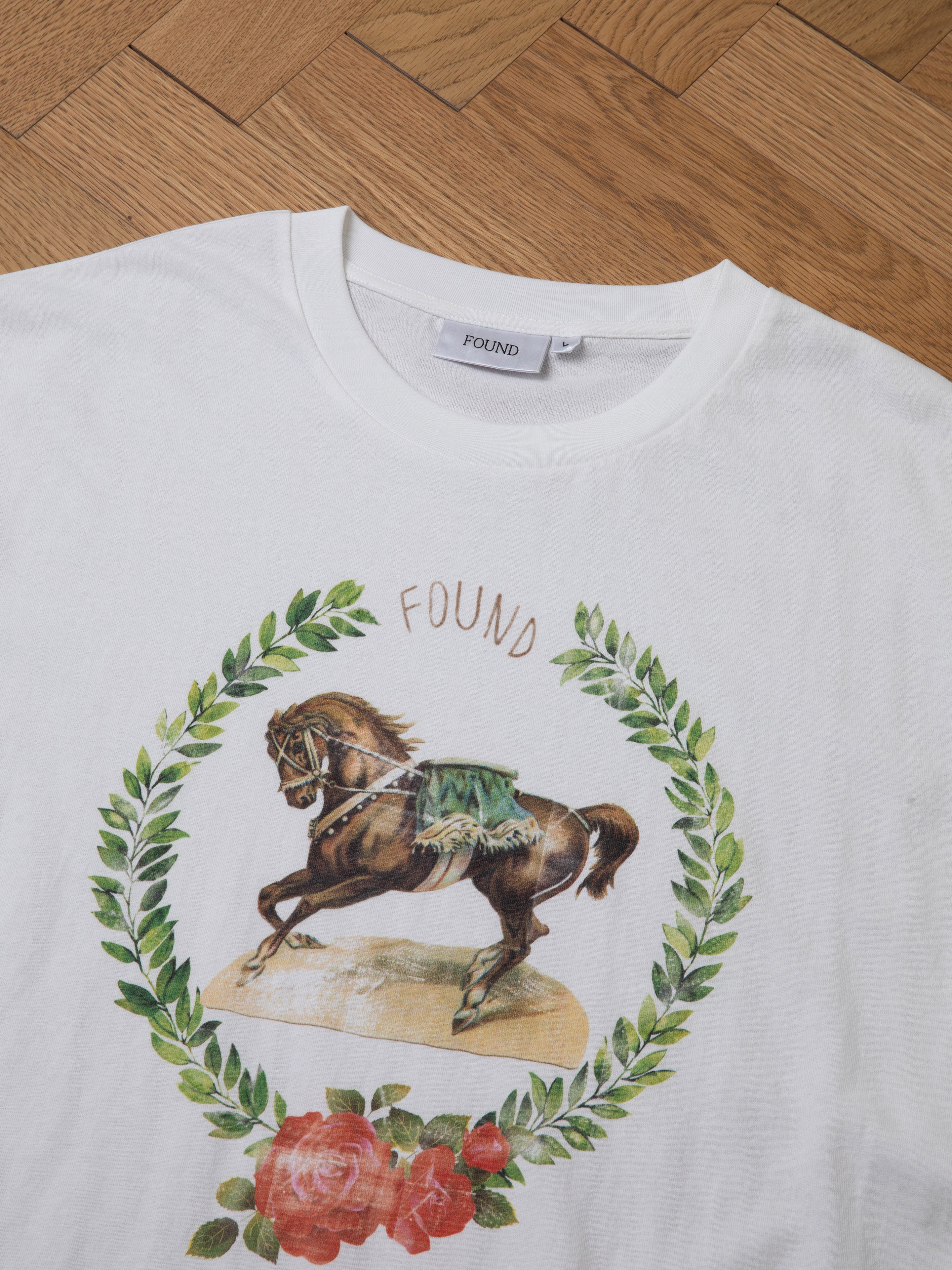 Introducing the Rodeo Tee by FOUND: This classic white T-shirt boasts a vintage aesthetic, showcasing a central illustration of a horse encircled by an ornate wreath of leaves and flowers, with the brand name "FOUND" prominently displayed above. Styled against a wooden floor background, this western graphic shirt exudes charm and nostalgia.