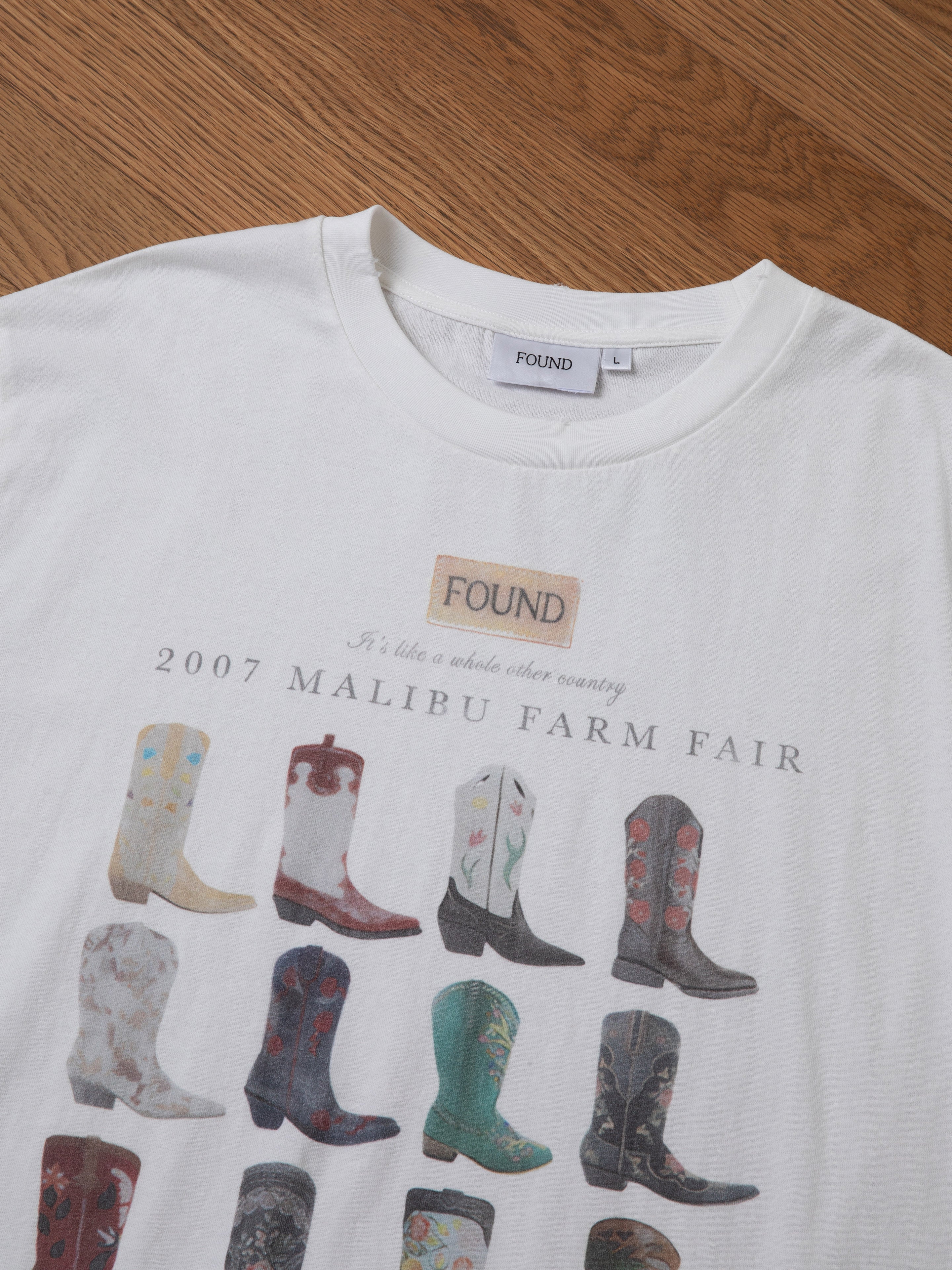 The Malibu Farm Fair Tee from FOUND is a 100% cotton white t-shirt featuring an illustrated design of various cowboy boots, with the text "2007 Malibu Farm Fair" printed on it. The label at the collar reads "FOUND." This vintage farm fair tee is displayed against a wooden surface background.