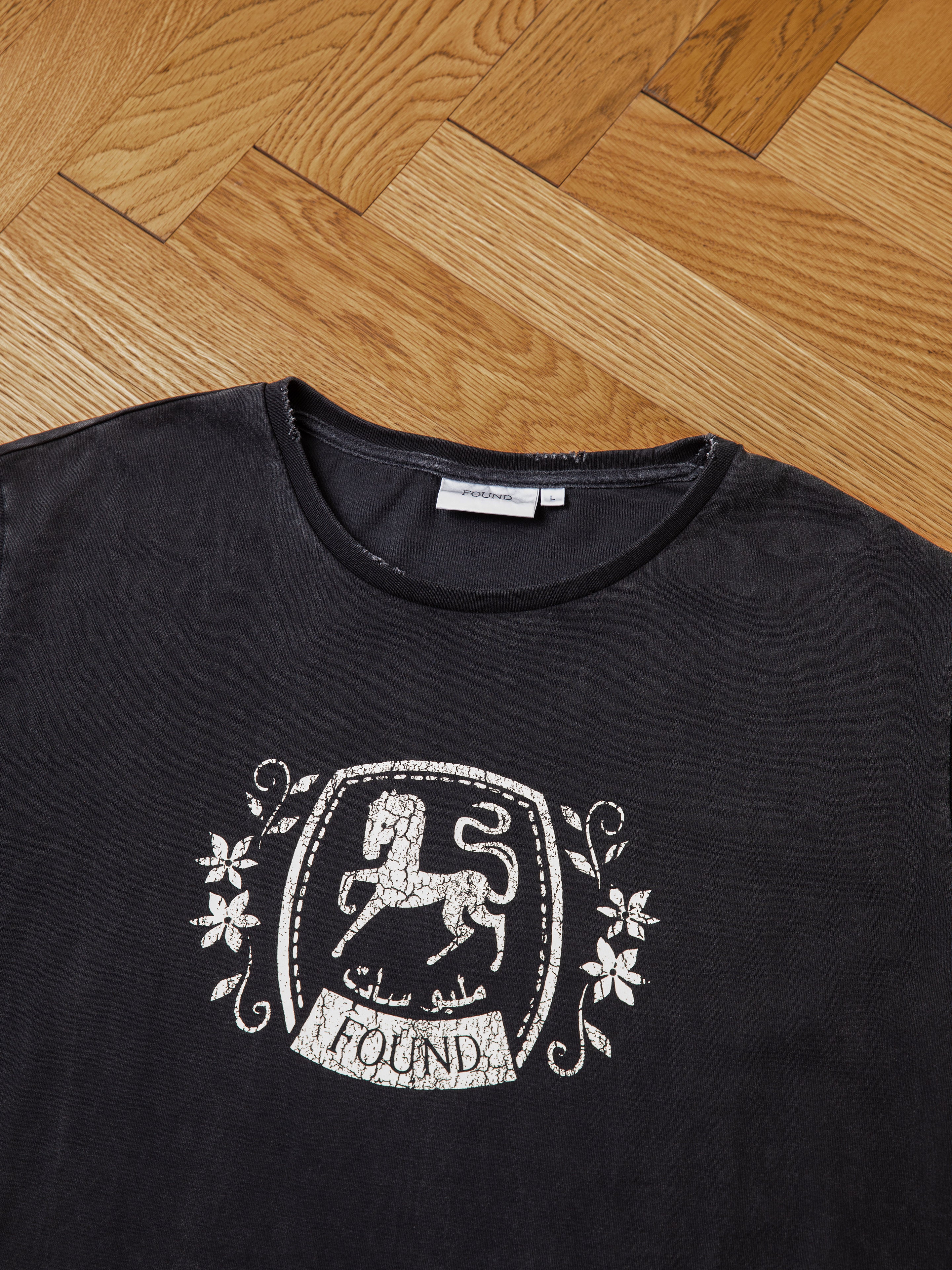 A black, 100% cotton Horse Logo Print Tee by FOUND, featuring a printed emblem of a lion with floral designs and the word "FOUND" in both English and Arabic. The shirt, enhanced by an enzyme wash for extra softness, is laid flat on a wooden herringbone-patterned floor.