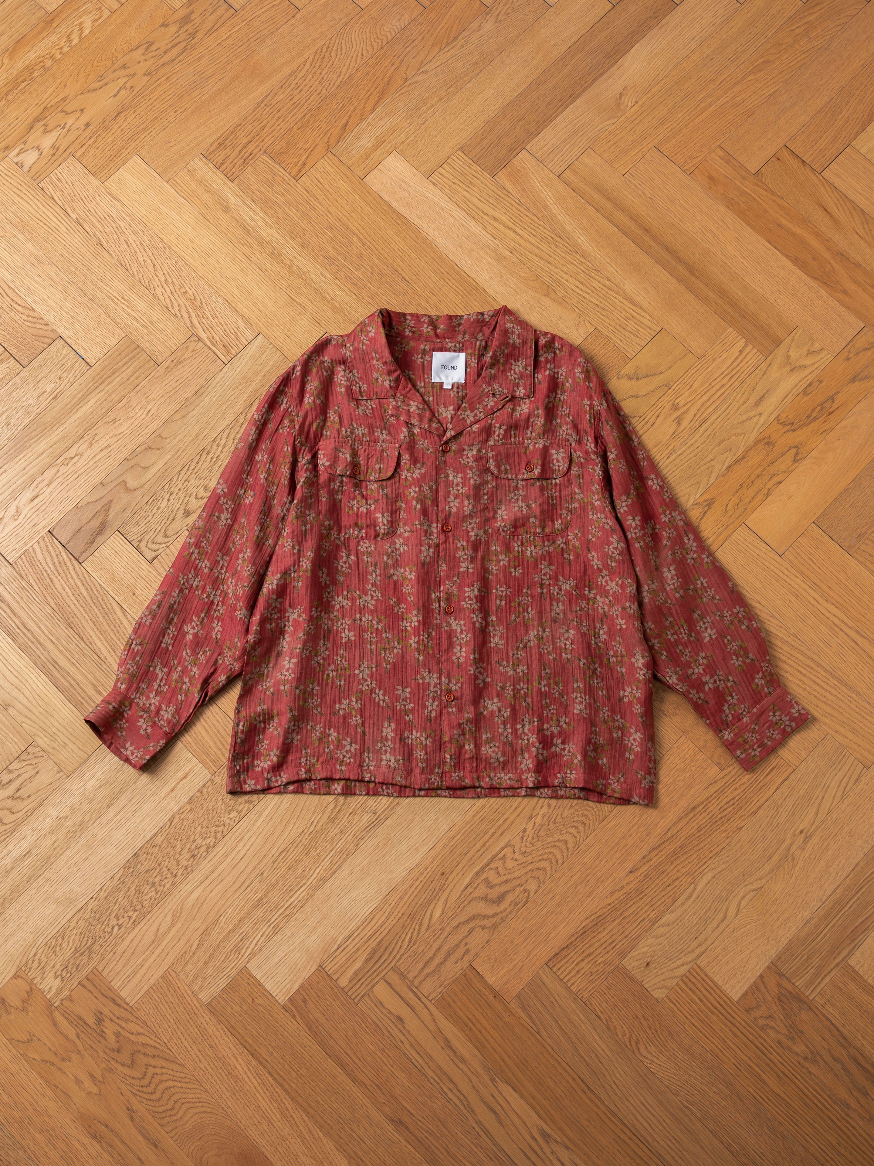 A Charlot LS Camp Shirt by FOUND, featuring a red floral pattern and dual chest pockets, is neatly laid out on a wooden herringbone-patterned floor.