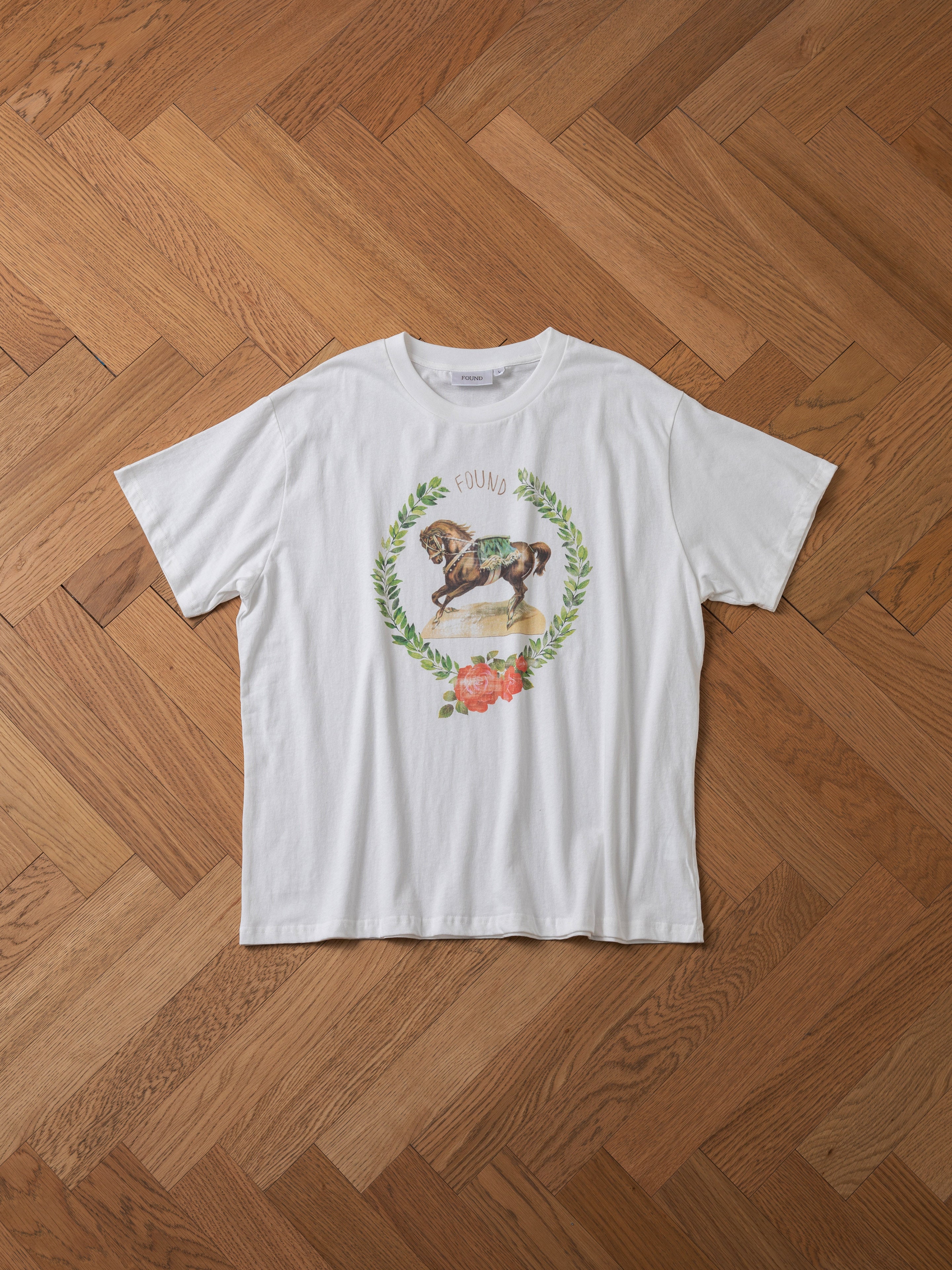 FOUND’s Rodeo Tee, showcasing a bird illustration within a laurel wreath on the chest, is set against a herringbone wooden floor and exudes a vintage feel.