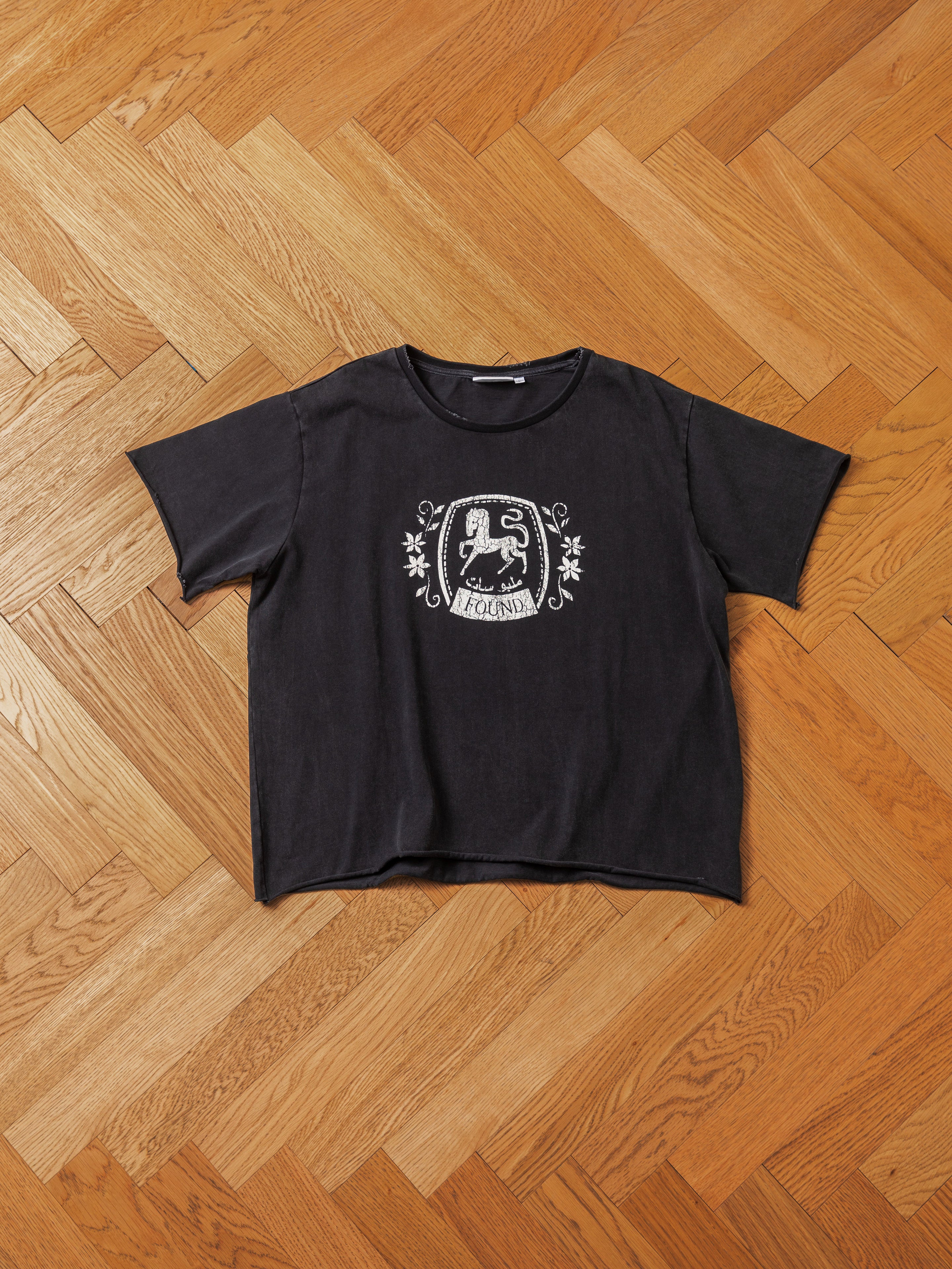 The Horse Logo Print Tee by FOUND, a black short-sleeve t-shirt featuring a lion emblem and decorative patterns, made from 100% cotton and treated with an enzyme wash, is laid out on a wooden herringbone floor.