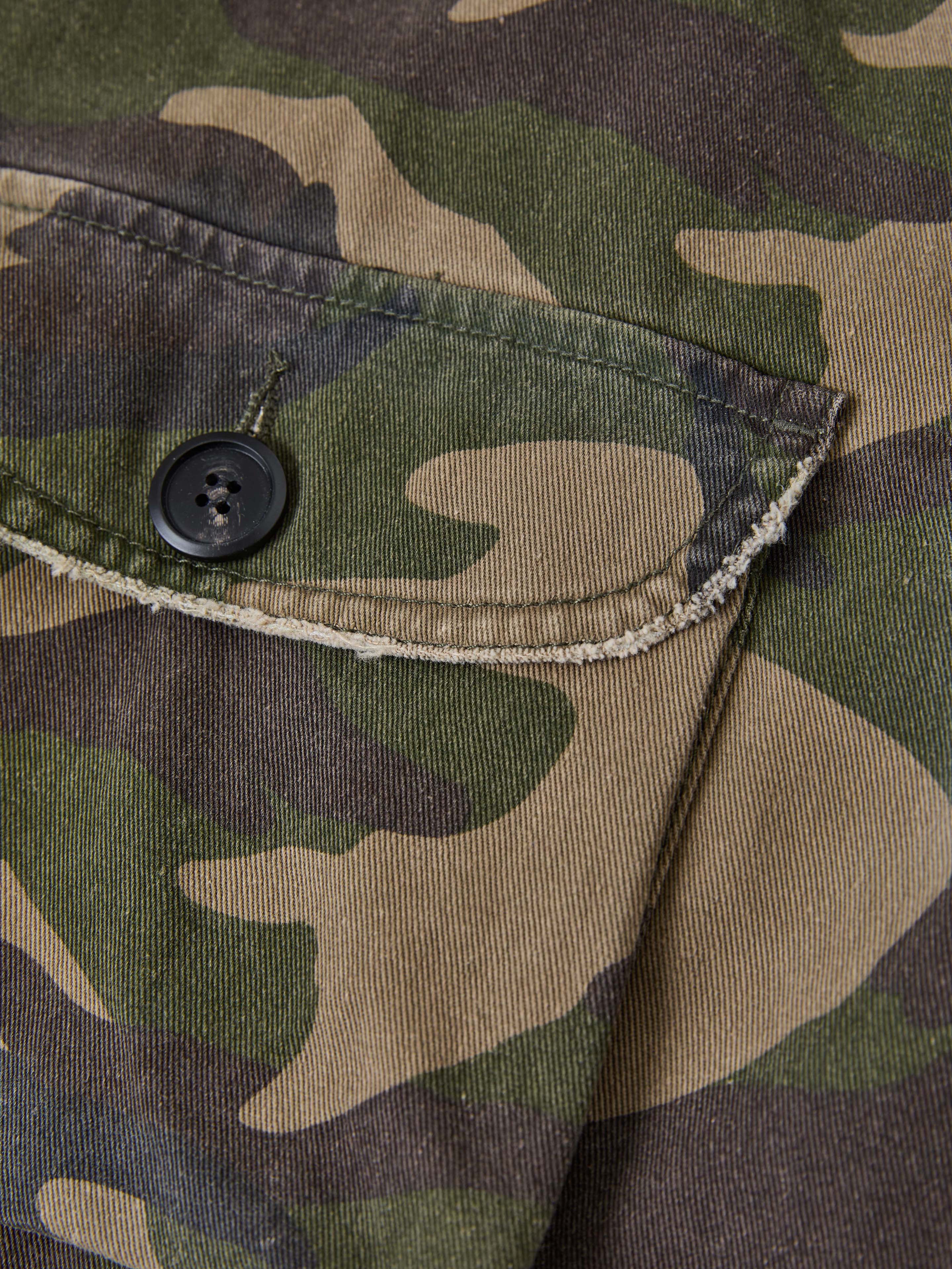 Close-up of a camo-patterned fabric with buttoned flap pockets, showcasing the vintage green, brown, and beige tones. The FOUND Camouflage Twill Utility Cargo Jacket emphasizes its rugged utility appeal through the visible seam and button details.