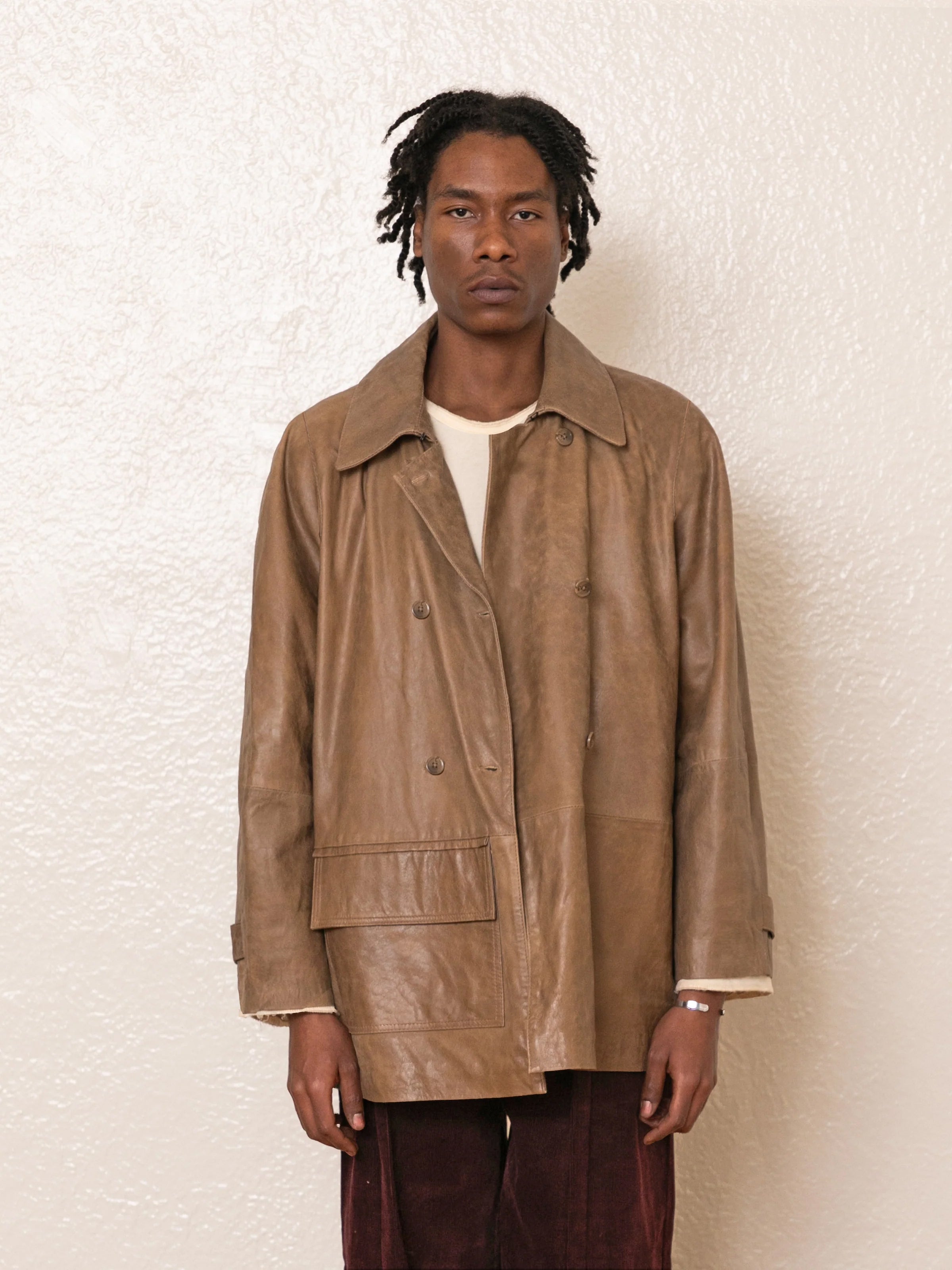 A person in a FOUND Fawn Double Breasted Leather Jacket over a white shirt stands against an off-white textured wall.