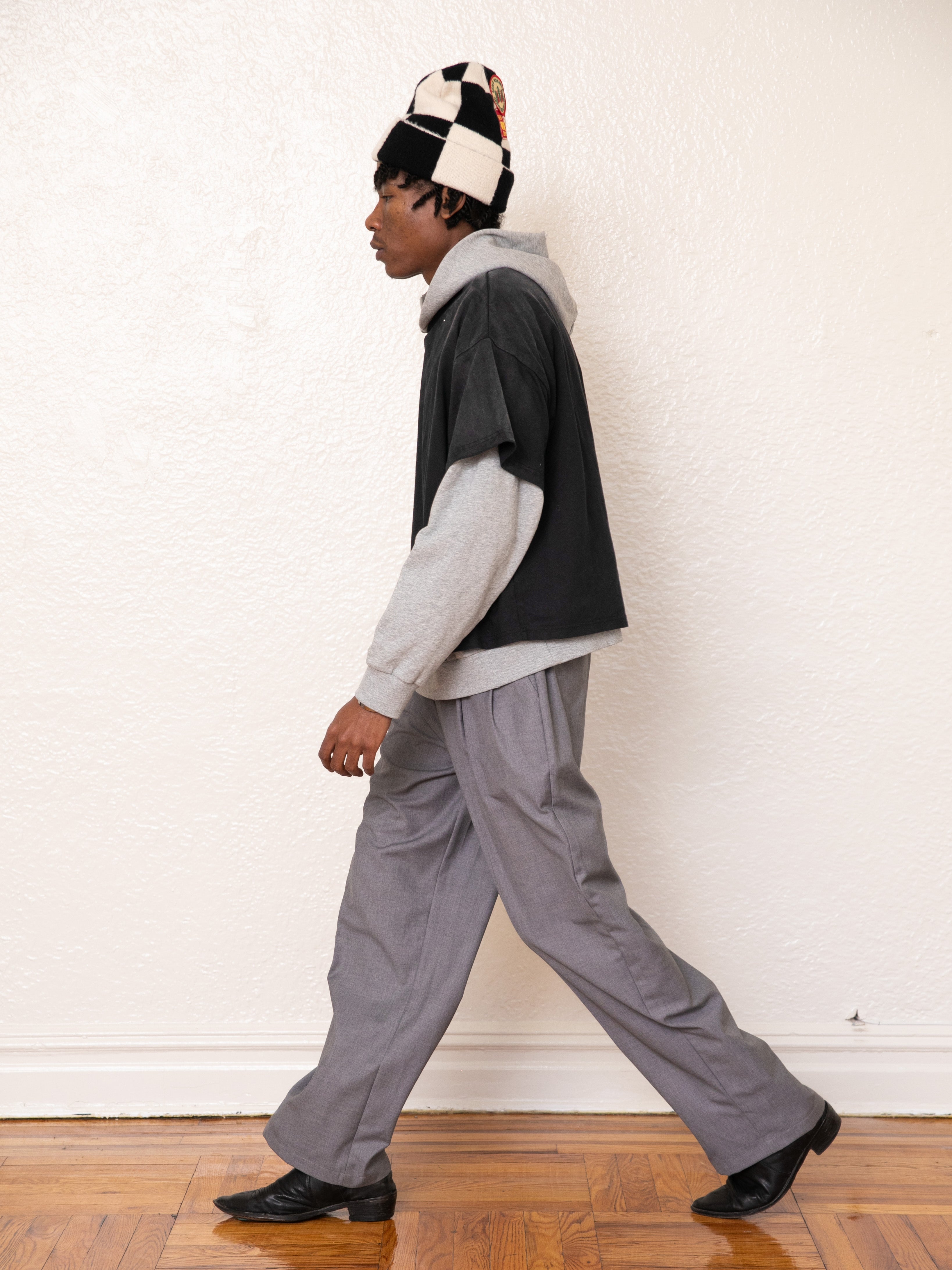 A person sporting a vintage look with a beanie, Found's Double Layer Hoodie in gray, a layered shirt, gray pants, and black shoes walks on a wooden floor indoors.