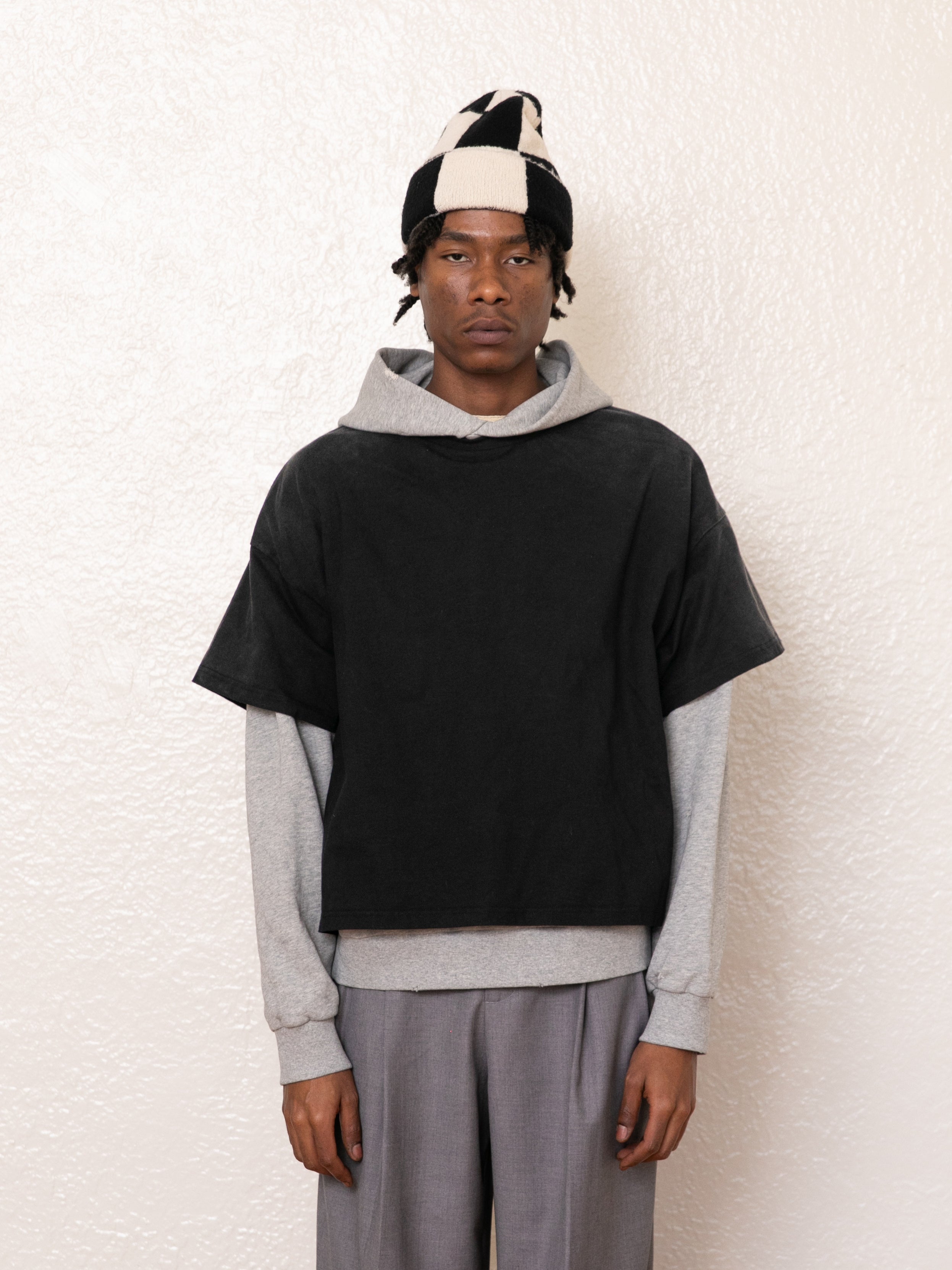 A person stands against a cream wall, wearing a geometric-patterned hat, black short-sleeve top, and gray pants. The vintage look is completed with the Double Layer Hoodie by Found, crafted from soft French terry cotton.