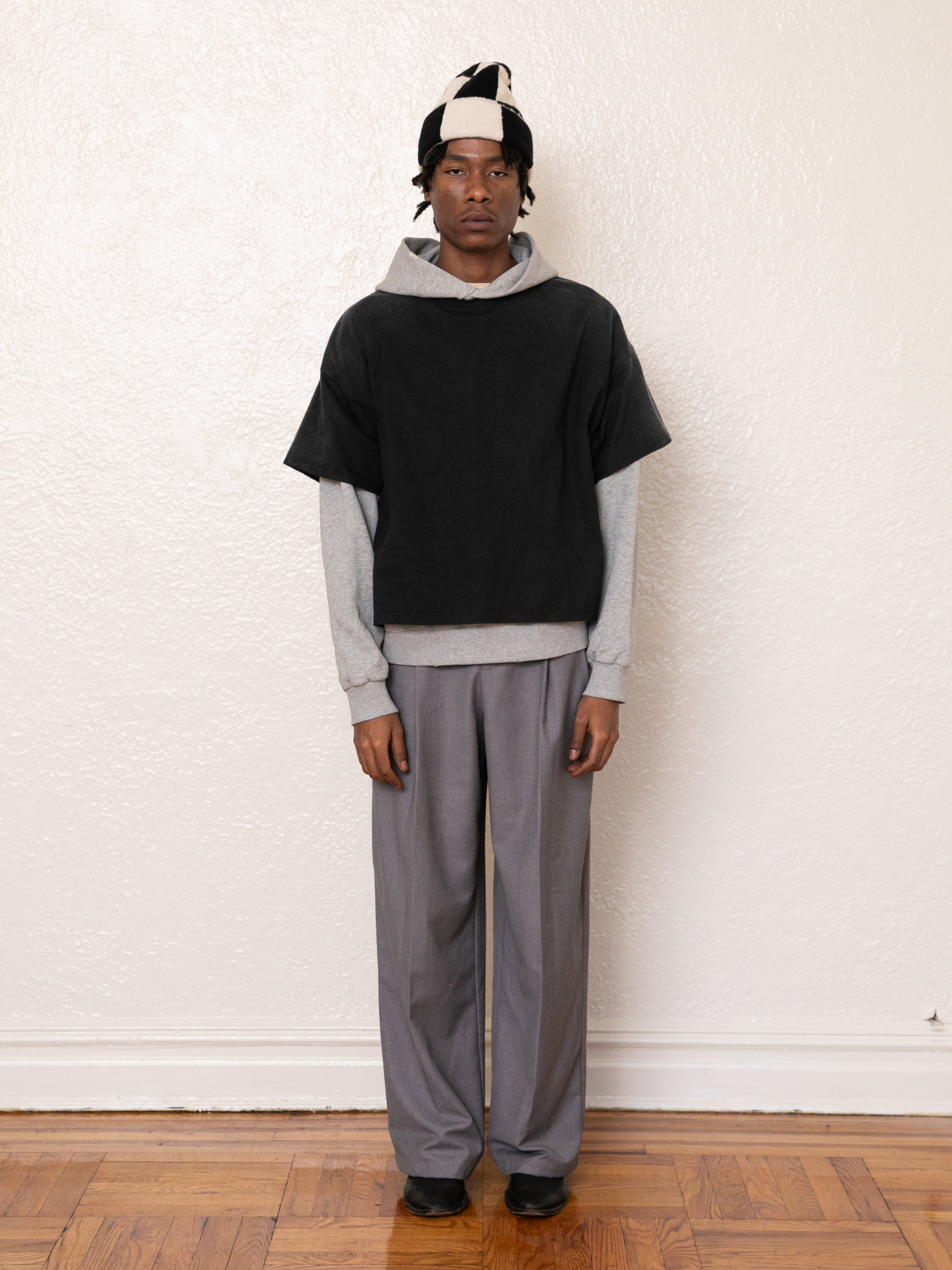 A person poses against a plain wall, wearing a vintage look featuring the Found Double Layer Hoodie of French terry cotton, paired with a black short-sleeve top, gray trousers, and a black-and-white beanie.