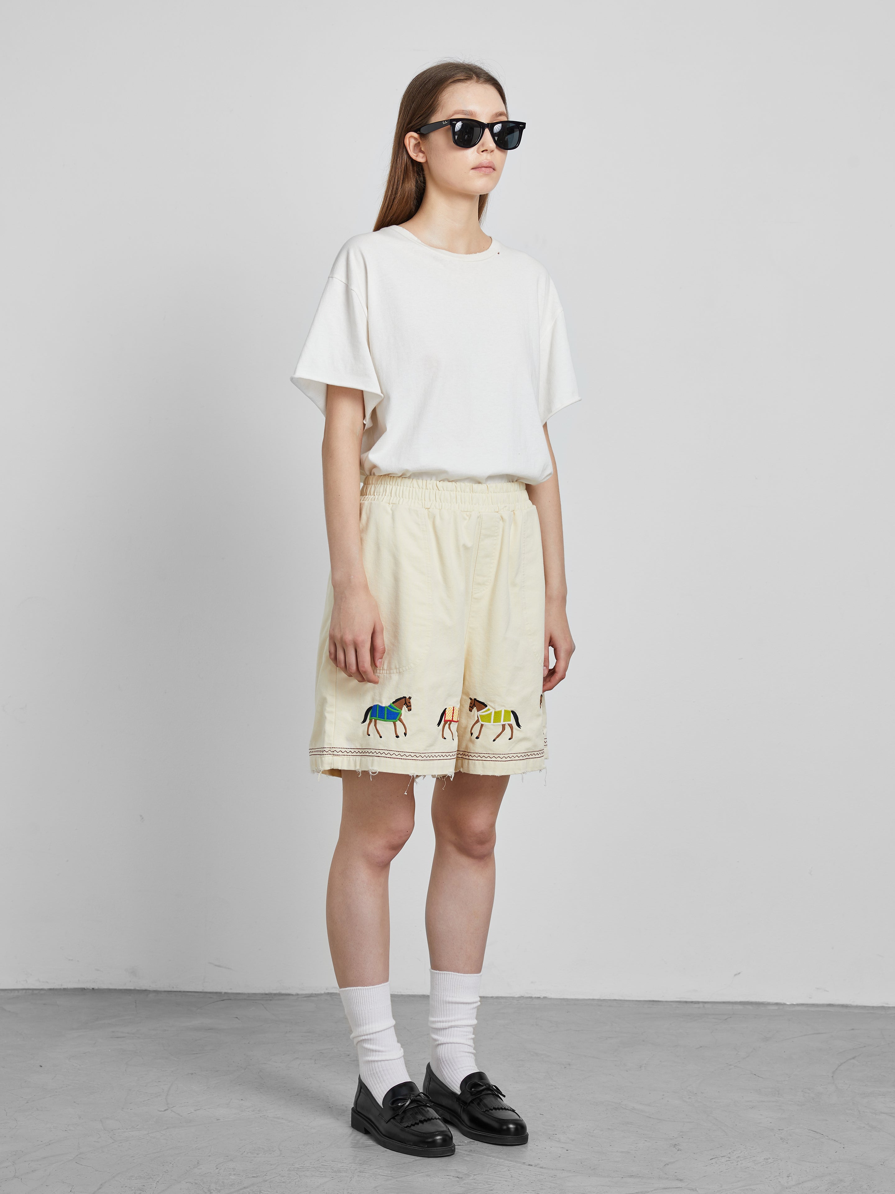 A person stands against a plain gray backdrop, donning sunglasses, a white t-shirt, and cream cotton twill shorts featuring embroidered horse motifs from Profound's Embroidered Horse Shorts collection.