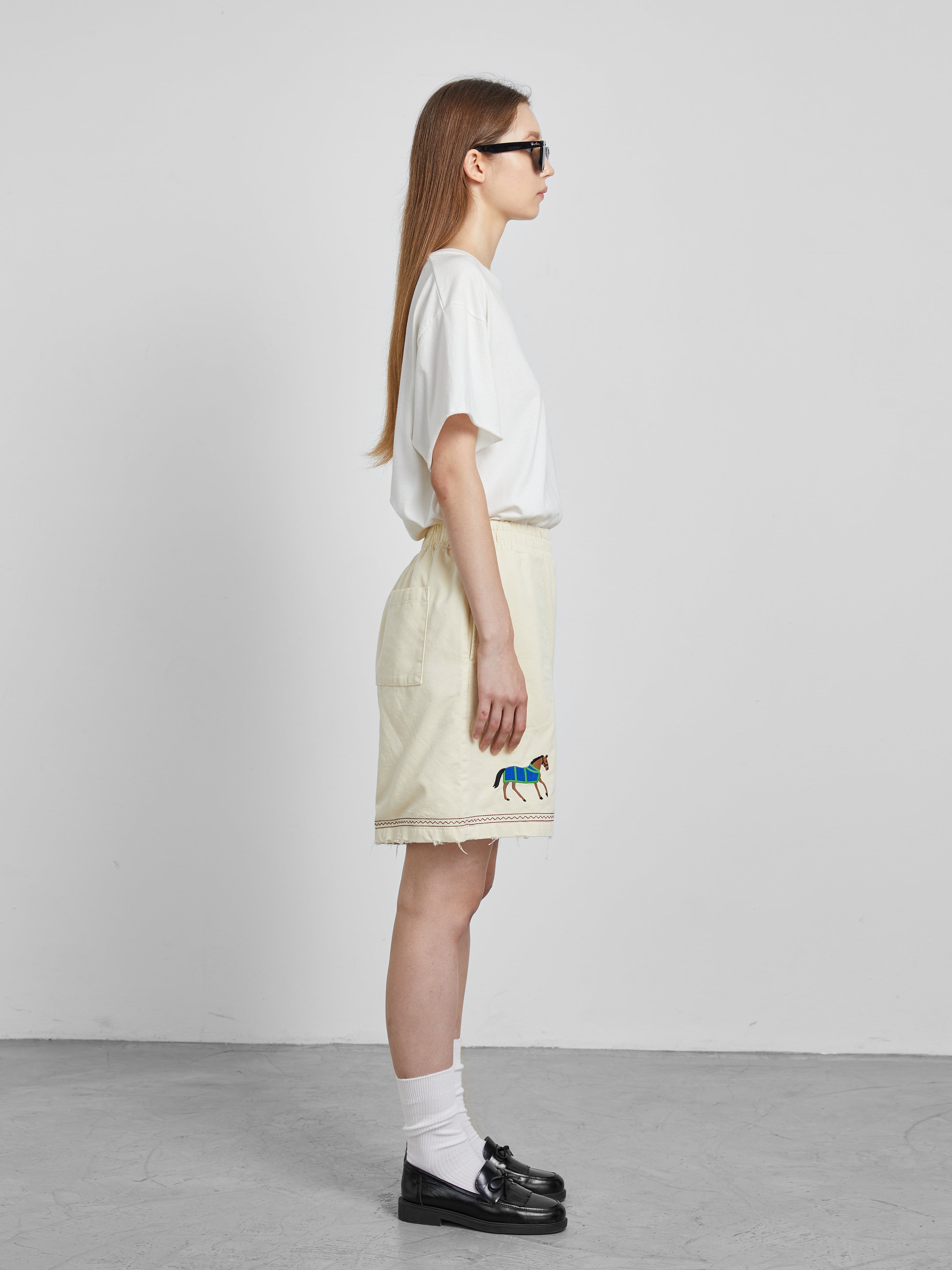 A woman stands in profile wearing a white t-shirt and cream-colored shorts from Profound, beautifully adorned with embroidered horse motifs. She completes her look with white socks, black loafers, sunglasses, and long flowing hair.