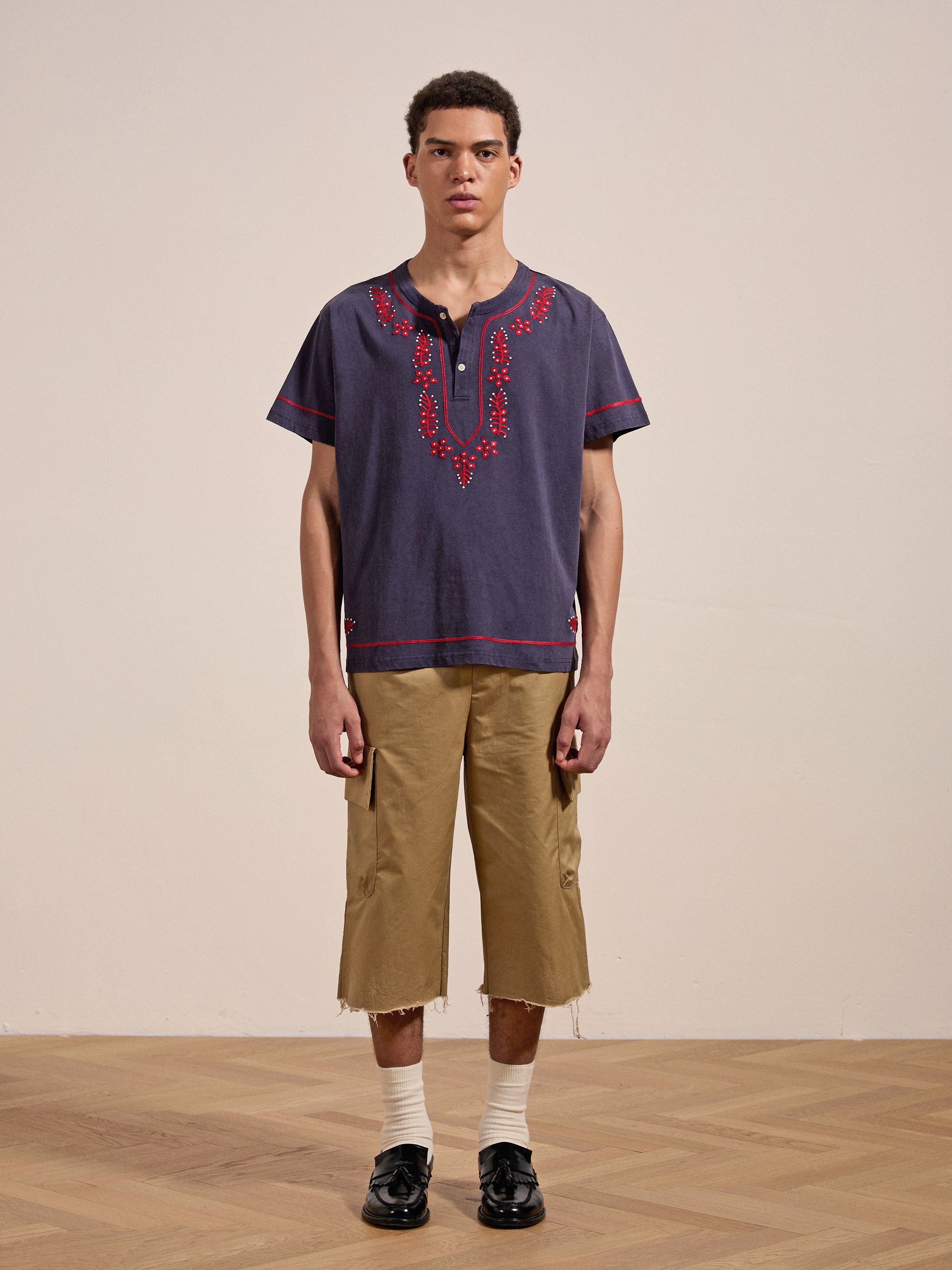 A person stands indoors on a wooden floor wearing the Embroidered Henley by FOUND, tan knee-length shorts, white socks, and black shoes, facing the camera.