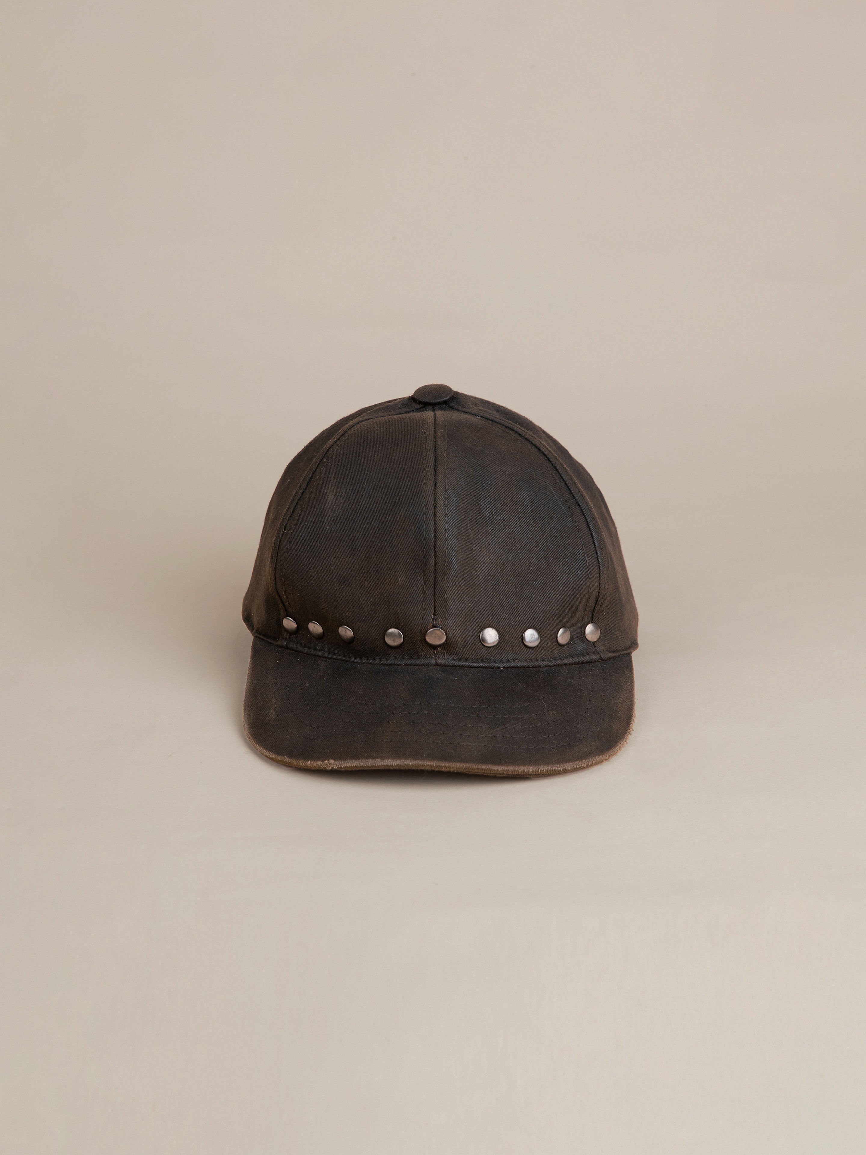 The Waxed Charcoal Studded Cap by FOUND features a worn, dark cotton canvas with antique matte silver studs along the front edge, displayed against a plain beige background.