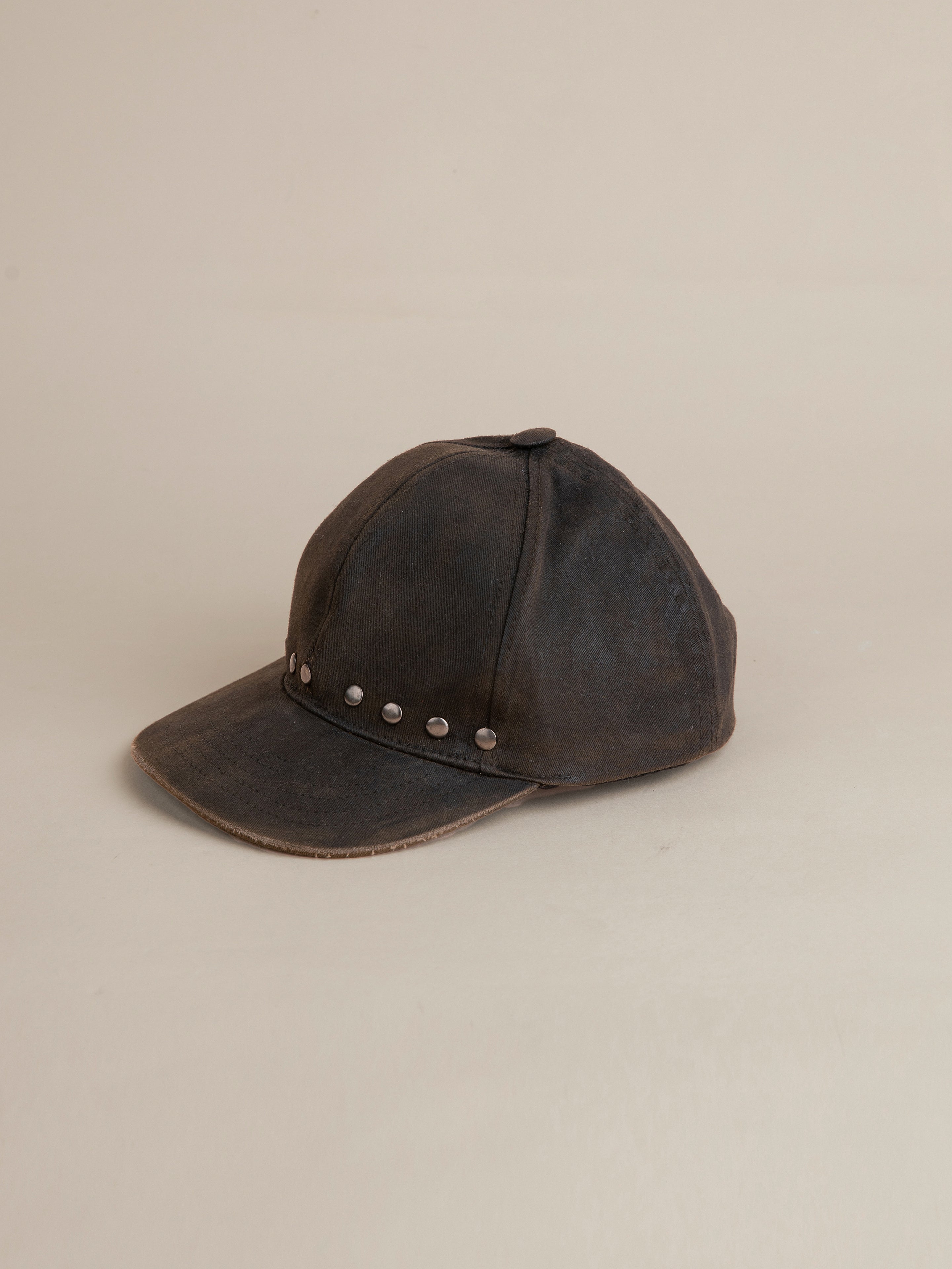 The Waxed Charcoal Studded Cap by FOUND, featuring antique matte silver studs lining the front, is placed on a light beige background.