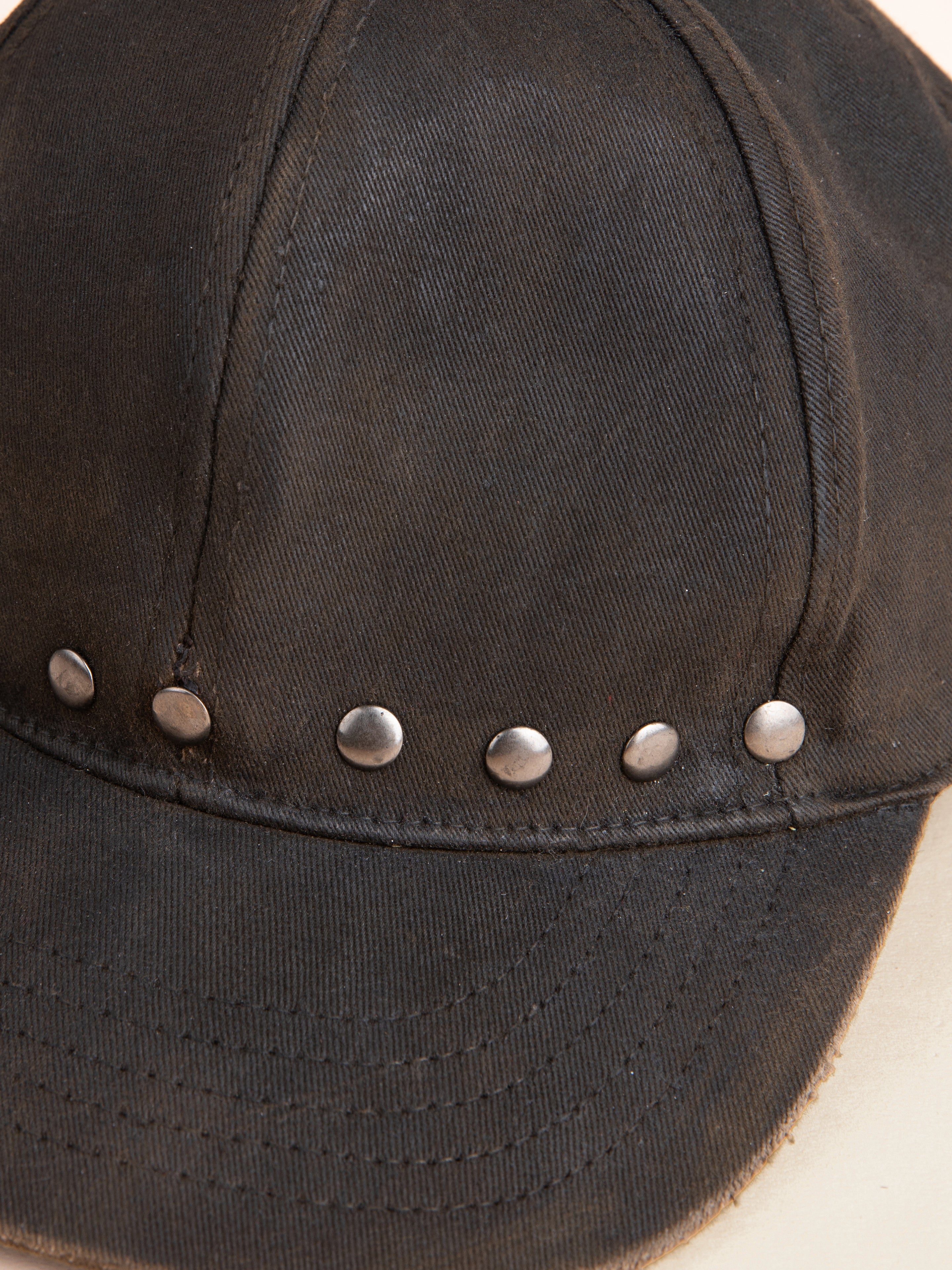 A close-up of the Waxed Charcoal Studded Cap by FOUND, crafted from cotton canvas and adorned with a row of antique matte silver studs along the front panel.