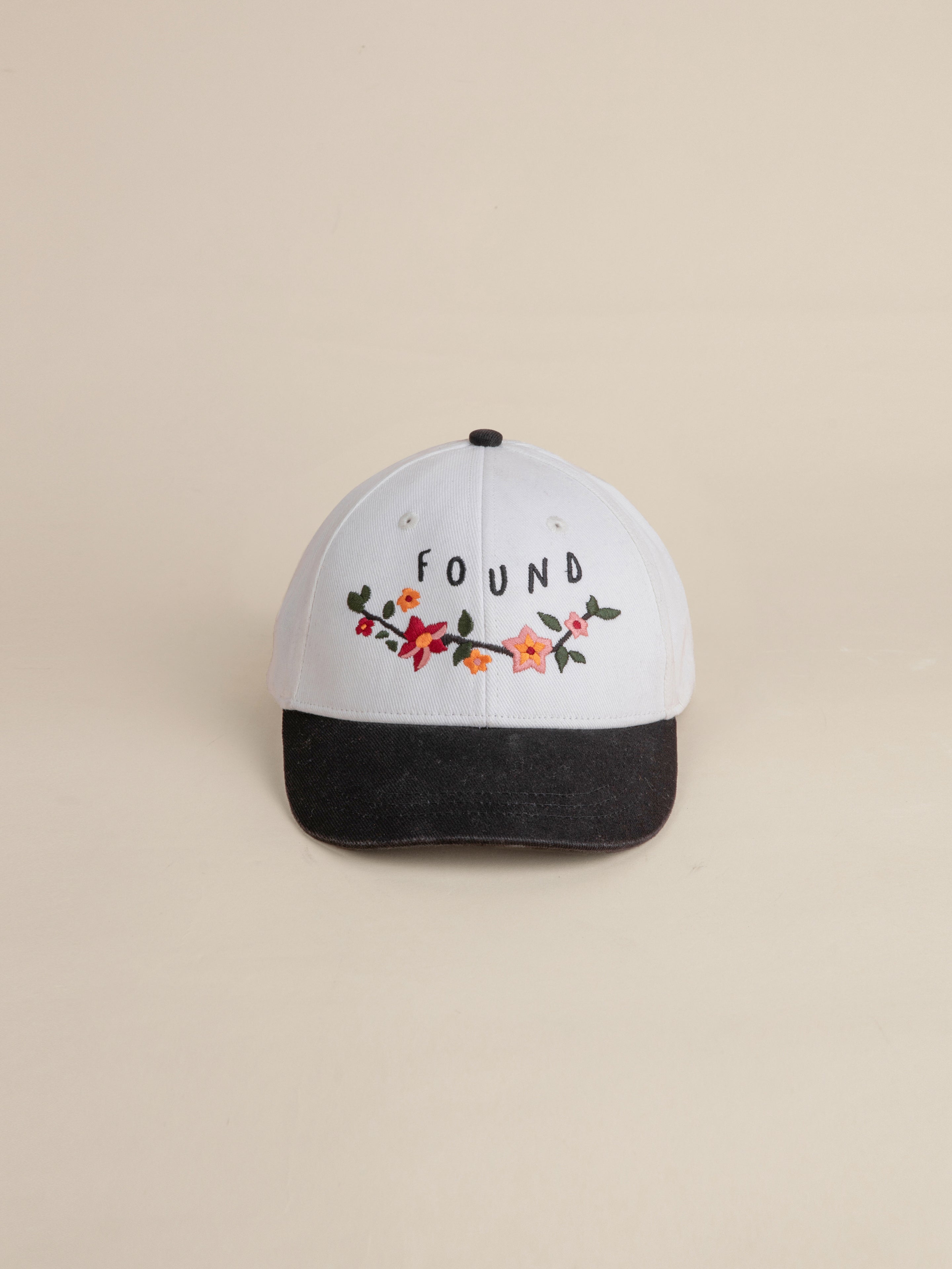 The Greenlands Contrast Cap by FOUND is a white 6-panel curved brim baseball cap, accentuated with a black brim, showcasing Kantha-style floral embroidery and the word "FOUND" on the front.