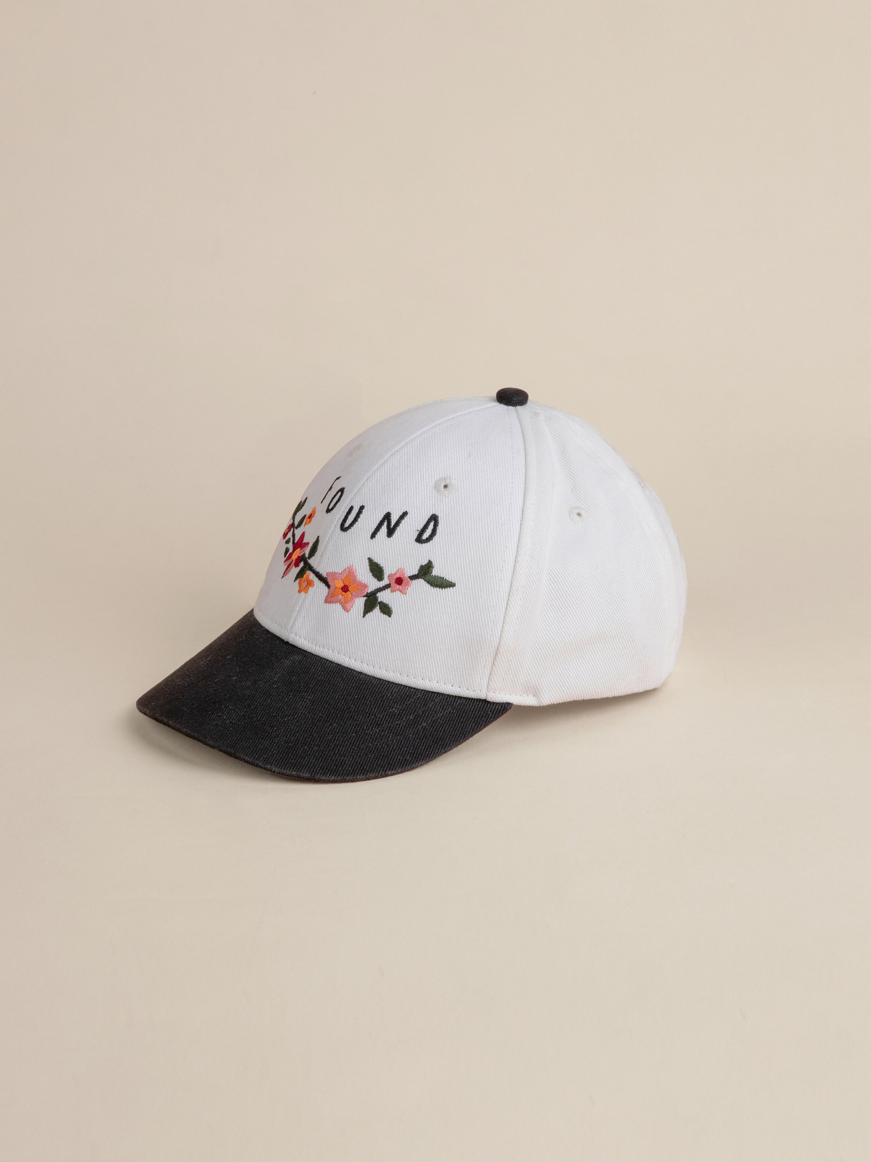 The Greenlands Contrast Cap by FOUND is a white cotton cap with a black visor, featuring Kantha-style floral embroidery and the brand name "FOUND" on the front.