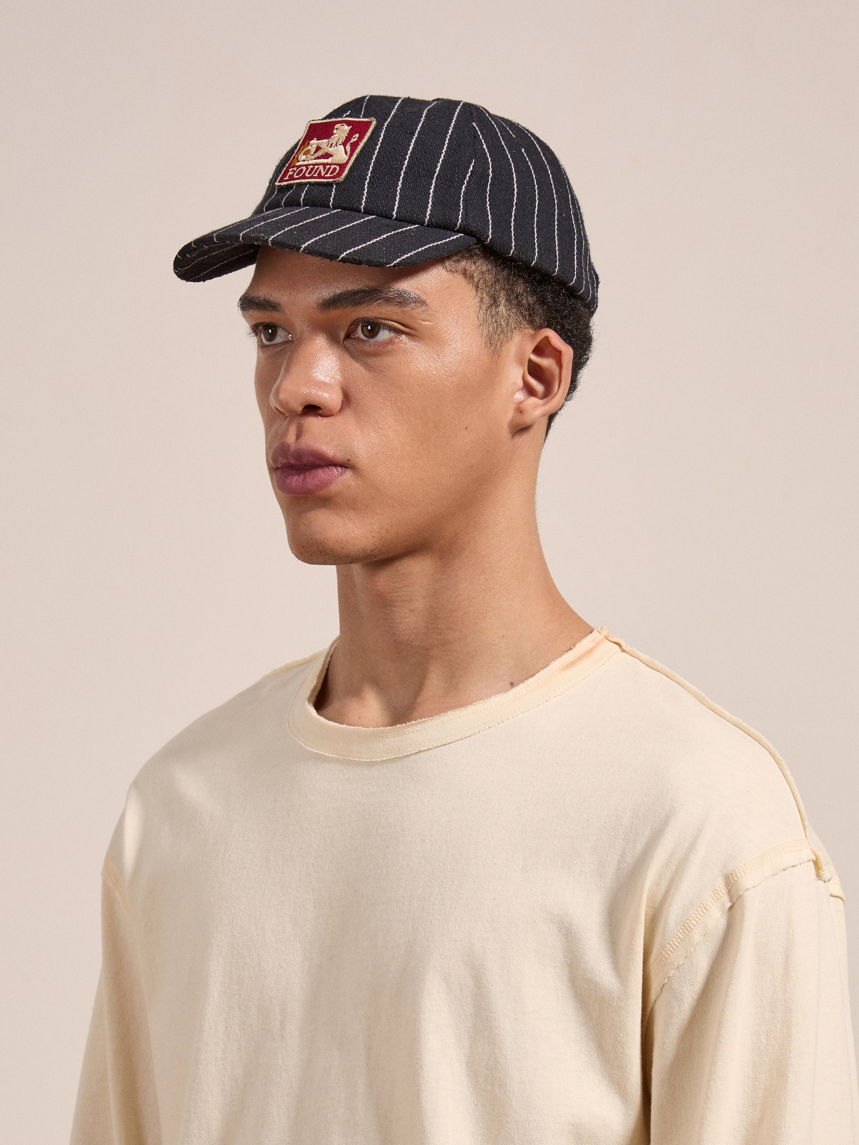 A person wearing a beige shirt and the FOUND Pinstripe Lion Cap featuring a red and beige patch is looking to the side with a neutral expression.