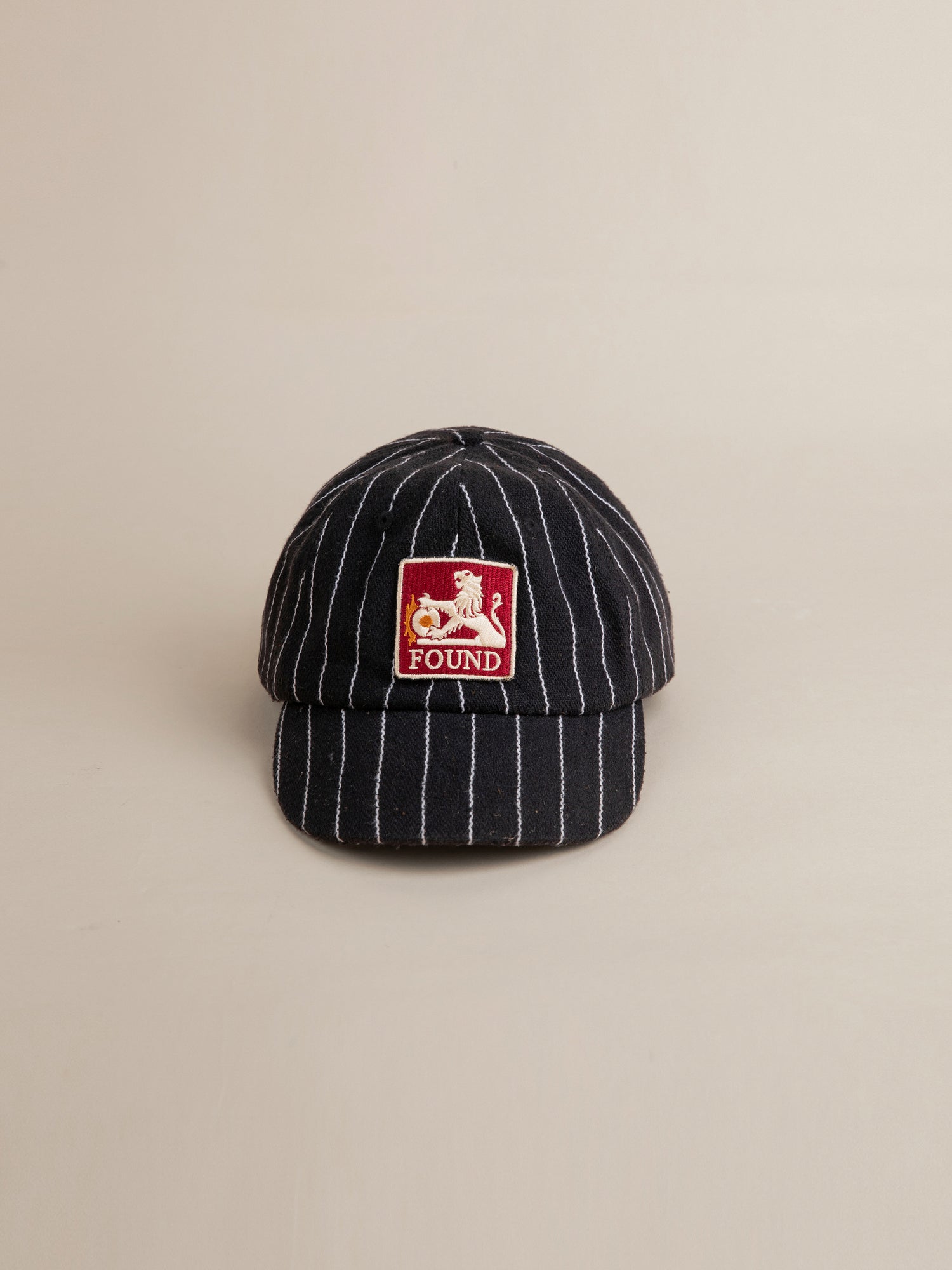 The Pinstripe Lion Cap from FOUND is a black, pinstriped baseball cap with a vintage wash and features a red and white embroidered patch reading 