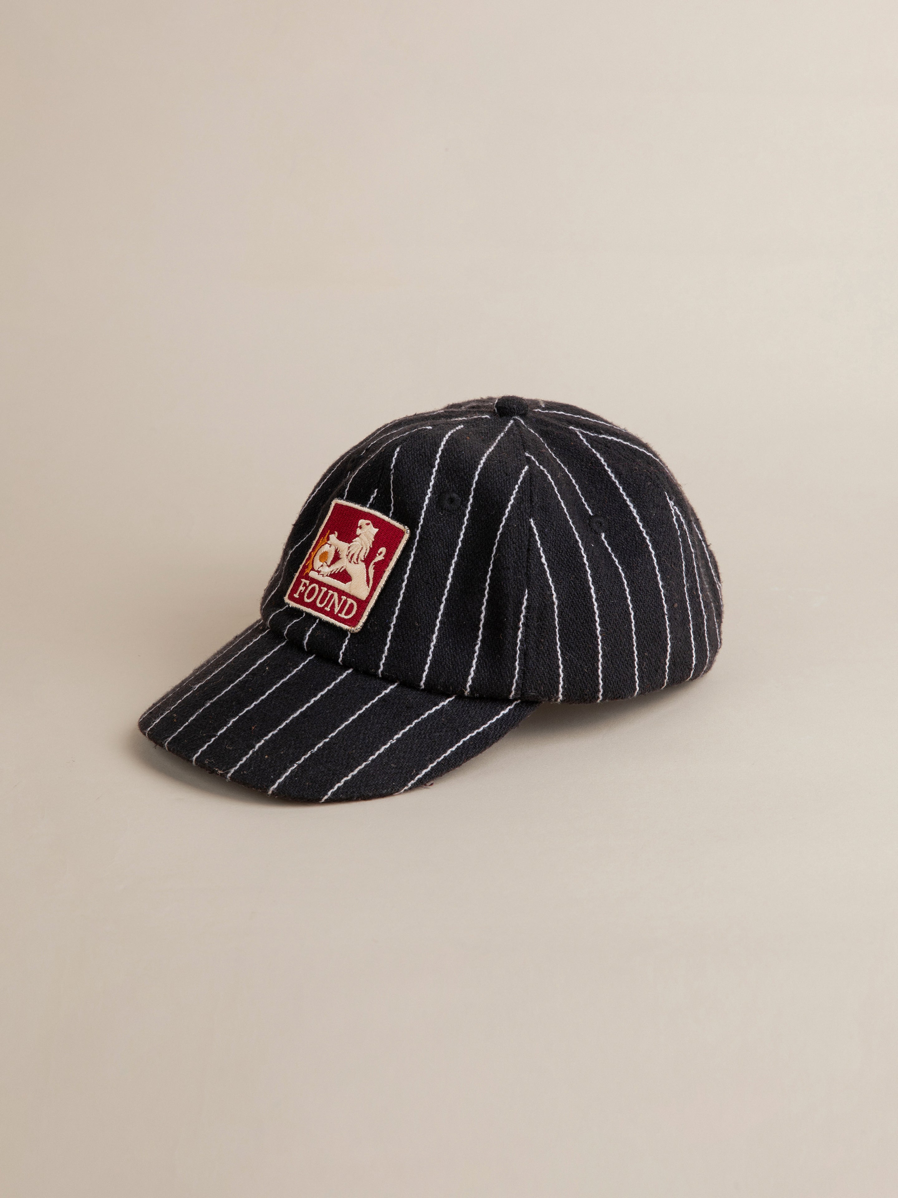 The Pinstripe Lion Cap by FOUND is a black baseball cap featuring white pinstripes along with a red patch on the front that displays an embroidered lion logo and the word "FOUND".