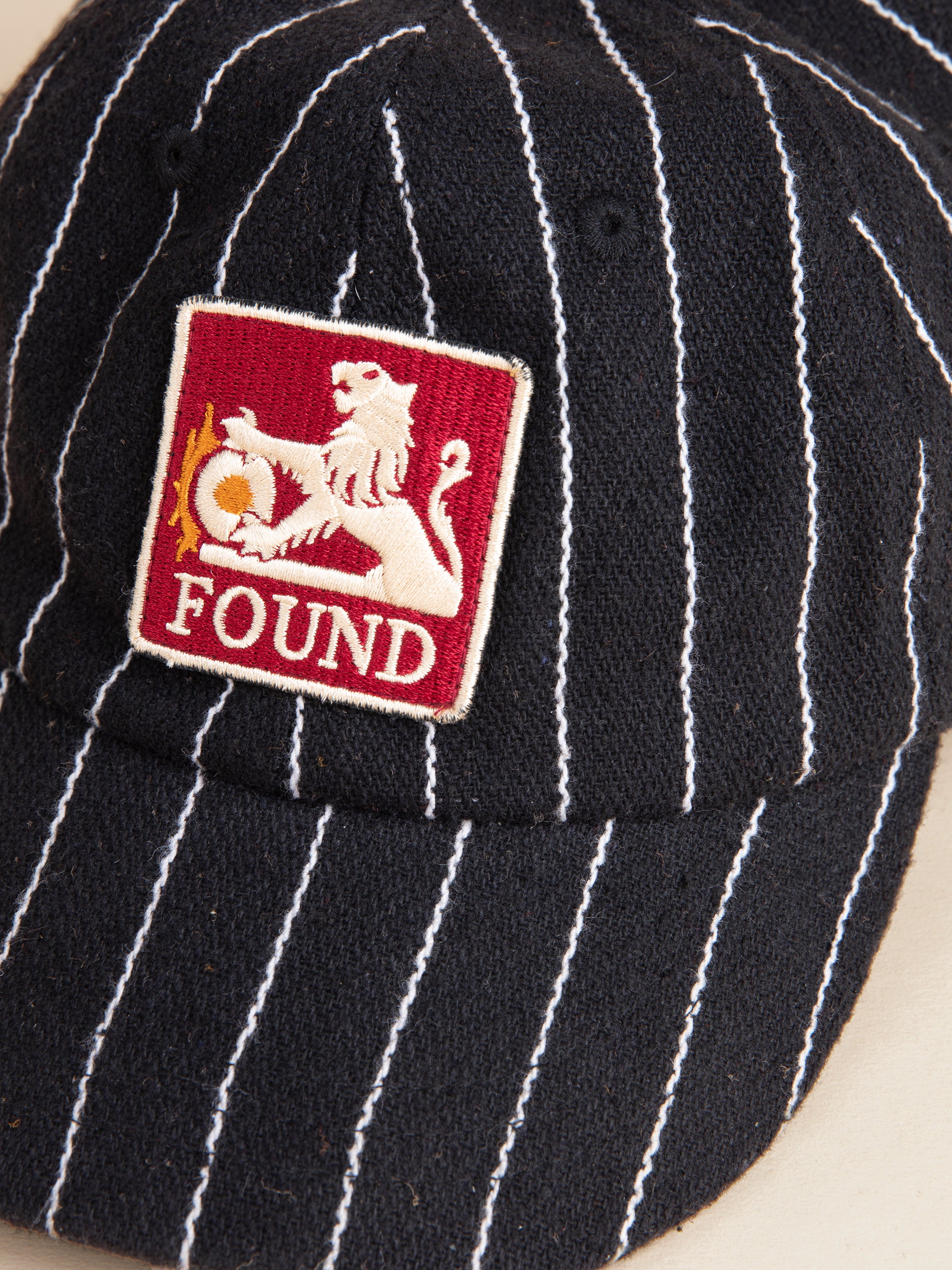 The Pinstripe Lion Cap from FOUND is a black, pinstriped design with an embroidered lion logo patch and the word "FOUND" artfully stitched on it.