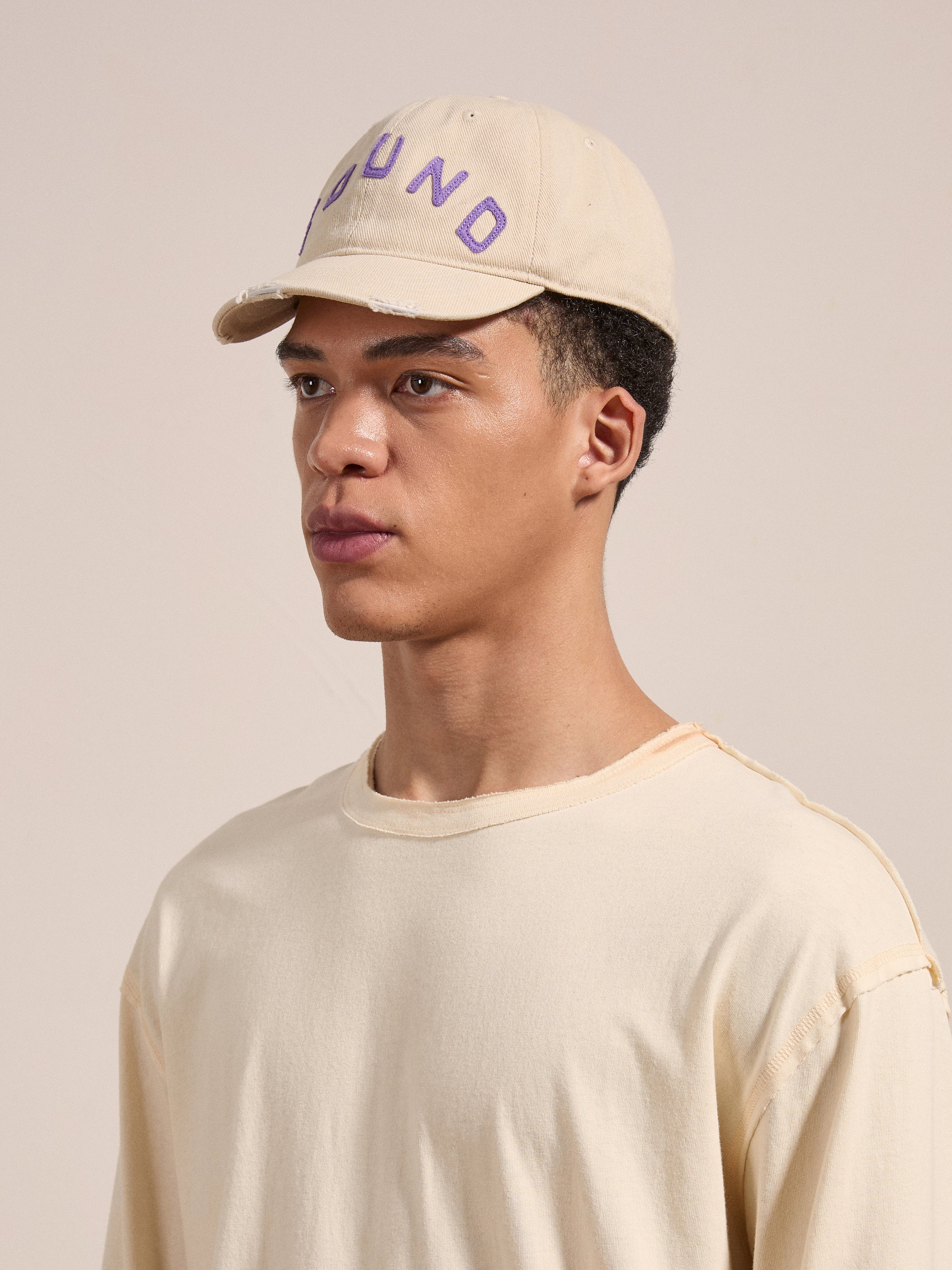 A person wearing an Ivory Felt Patch Cap from the brand FOUND and a beige shirt looks to the left against a plain background.