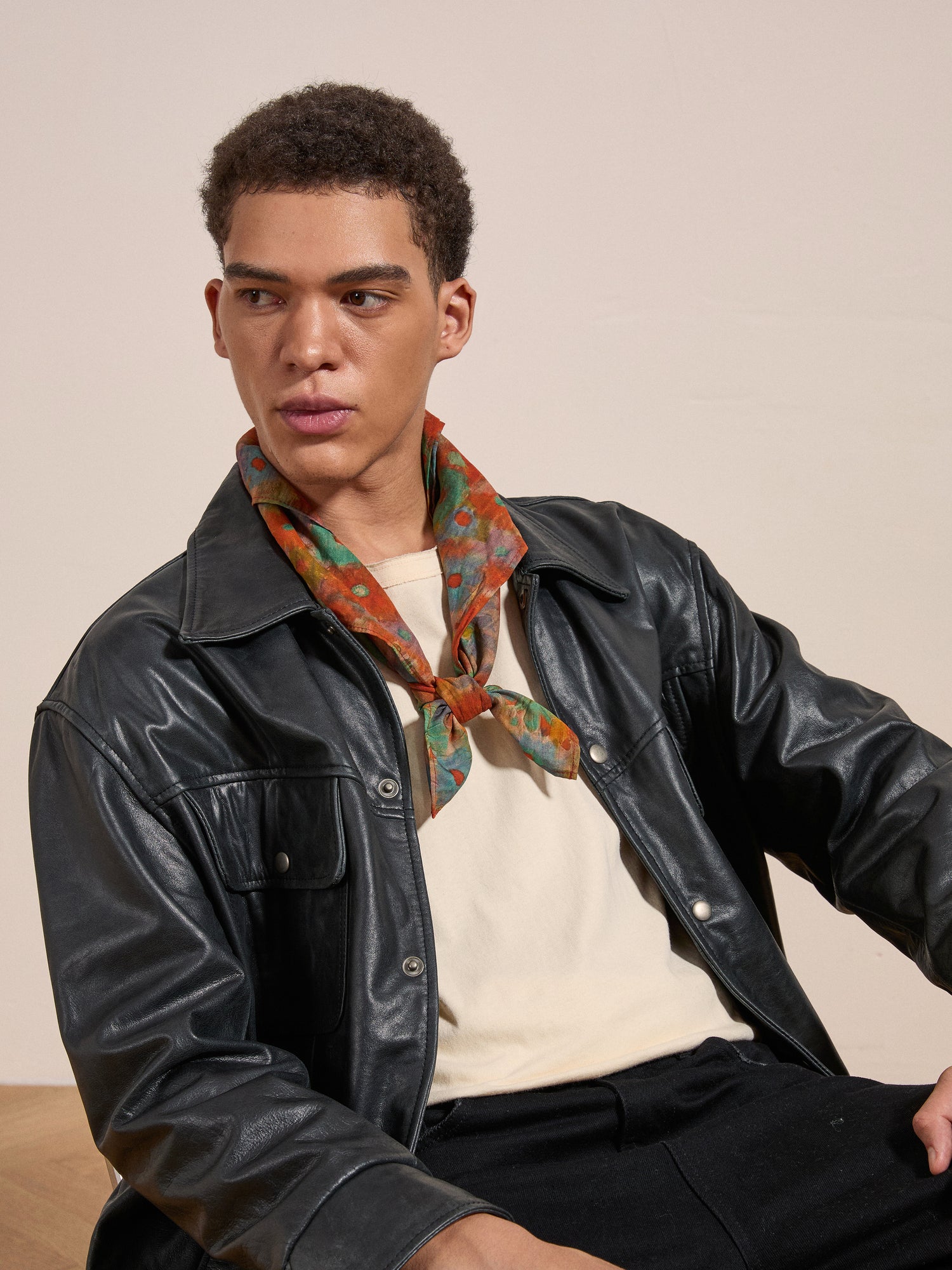 A person with short curly hair wears a black leather jacket, a white shirt, and the vibrant Sepehr Bandana by FOUND around their neck, sitting and looking off to the side.